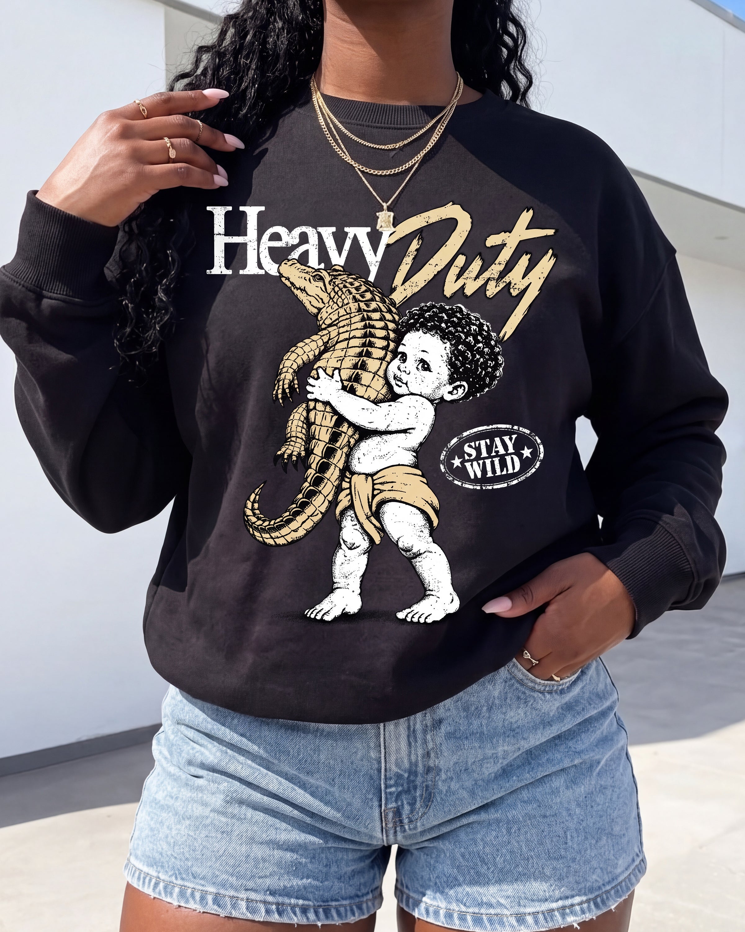 A woman wearing a black crewneck sweatshirt with a graphic print of a child holding an alligator, featuring the text 'Heavy Duty' and 'Stay Wild'.