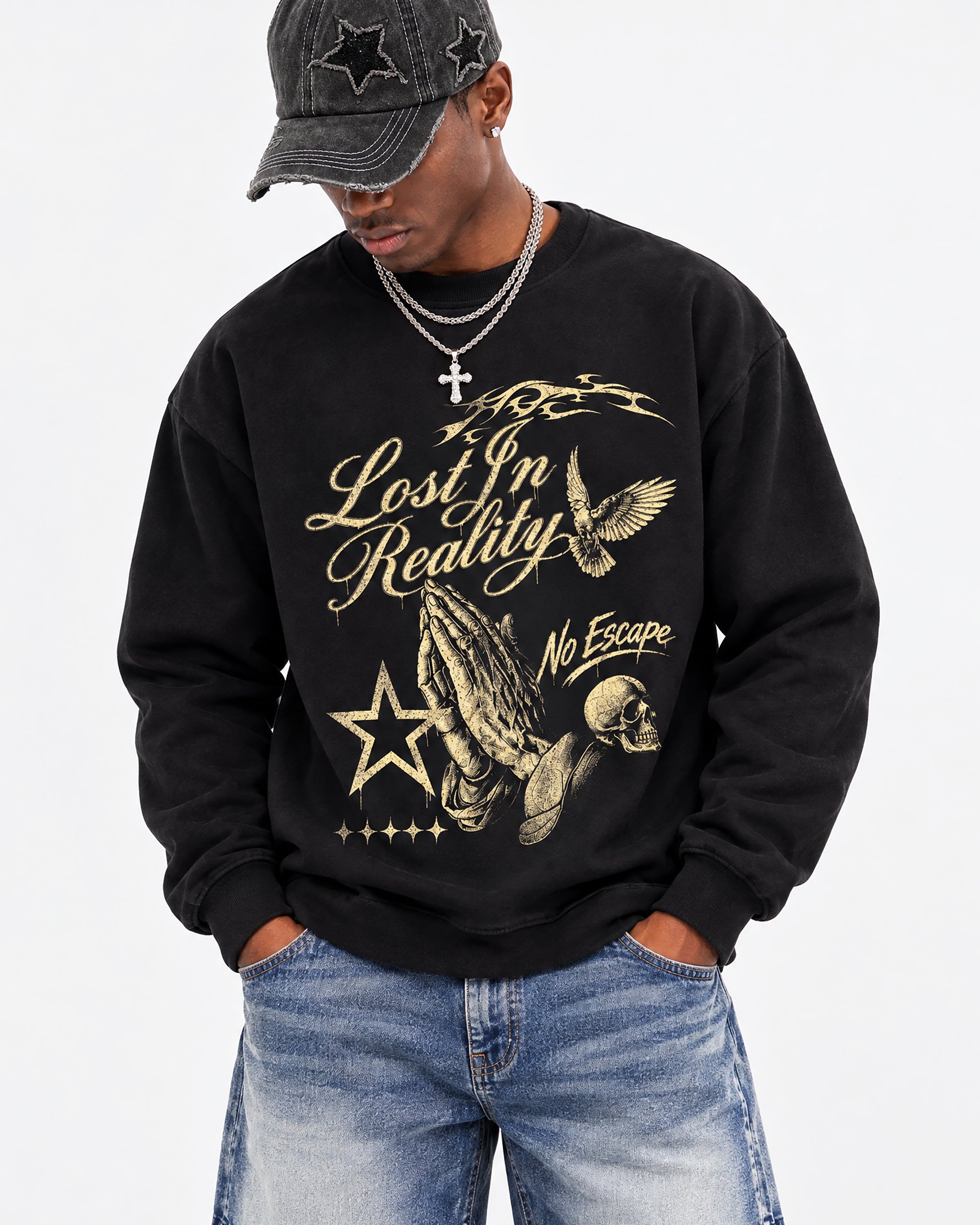 A man wearing a black crewneck sweatshirt with a gold graphic print that reads 'Lost In Reality No Escape' alongside a praying man, skull, and star design. He is also wearing a black star-studded baseball cap and blue jeans against a plain white background.