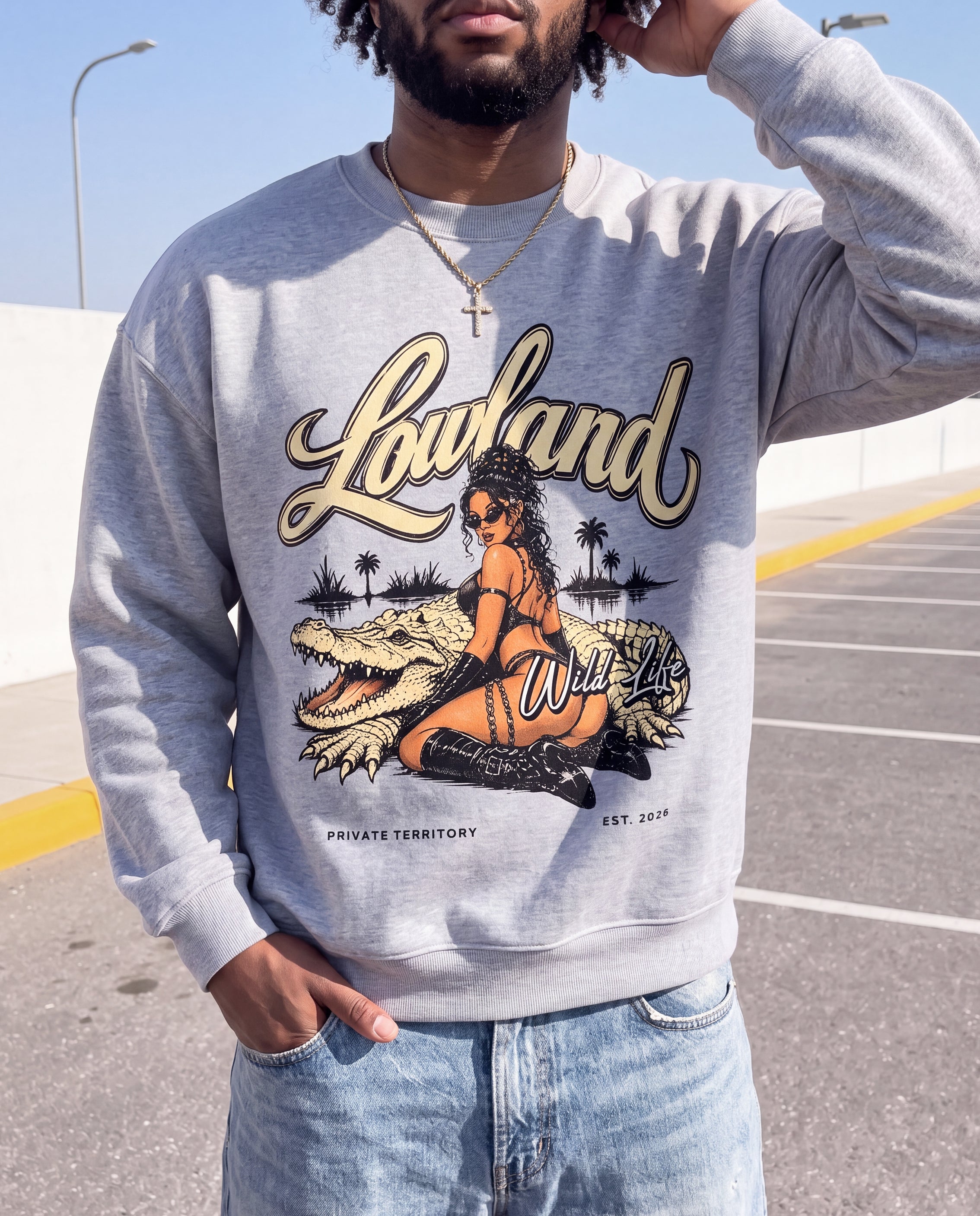A man wearing a heather gray crewneck sweatshirt with a graphic print of a woman riding an alligator, featuring the text 'Lowland Wild Life' and 'PRIVATE TERRITORY EST. 2019', paired with light blue jeans in an outdoor parking lot setting.