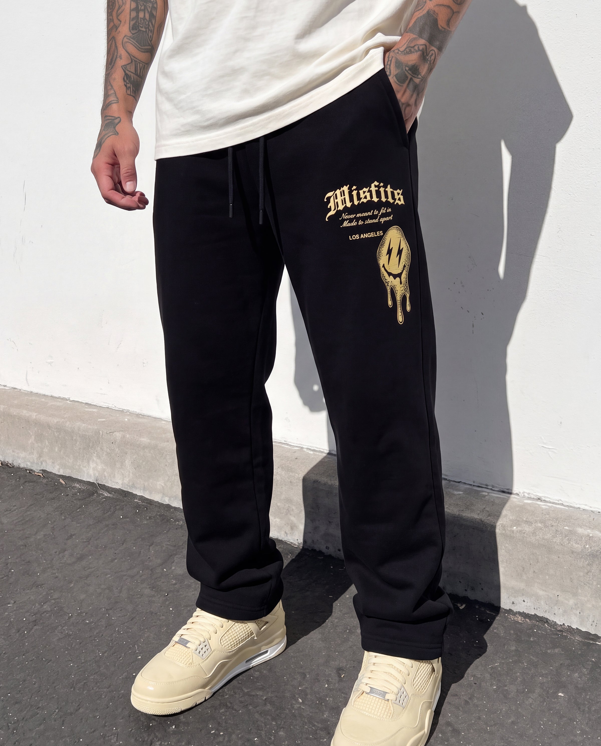 A person wearing black sweatpants with a gold Misfits logo and drip graphic on the left thigh, paired with a white t-shirt and beige sneakers, standing against a white wall.