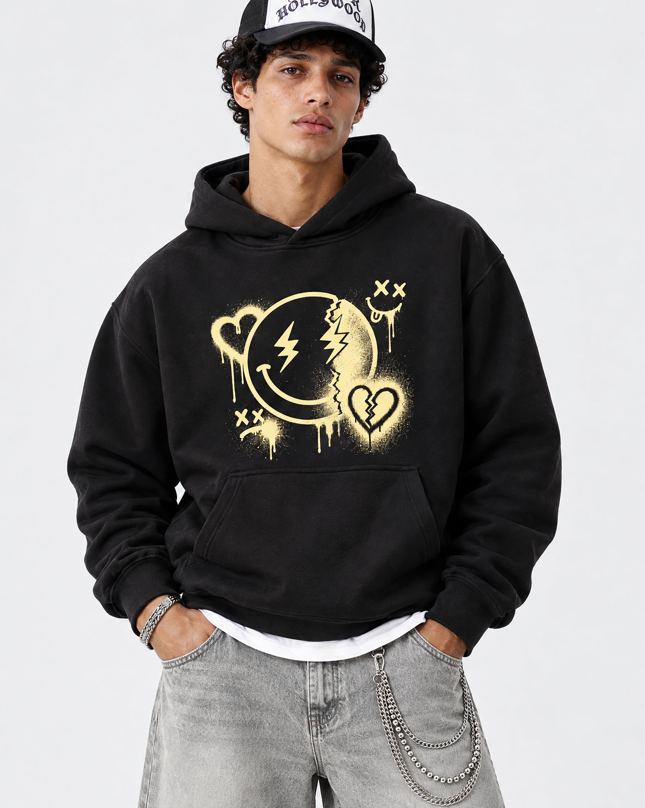 A man wearing a black pullover hoodie with a gold graphic print of a cracked moon, hearts, and 'XX' symbols, paired with light gray jeans and a black cap.