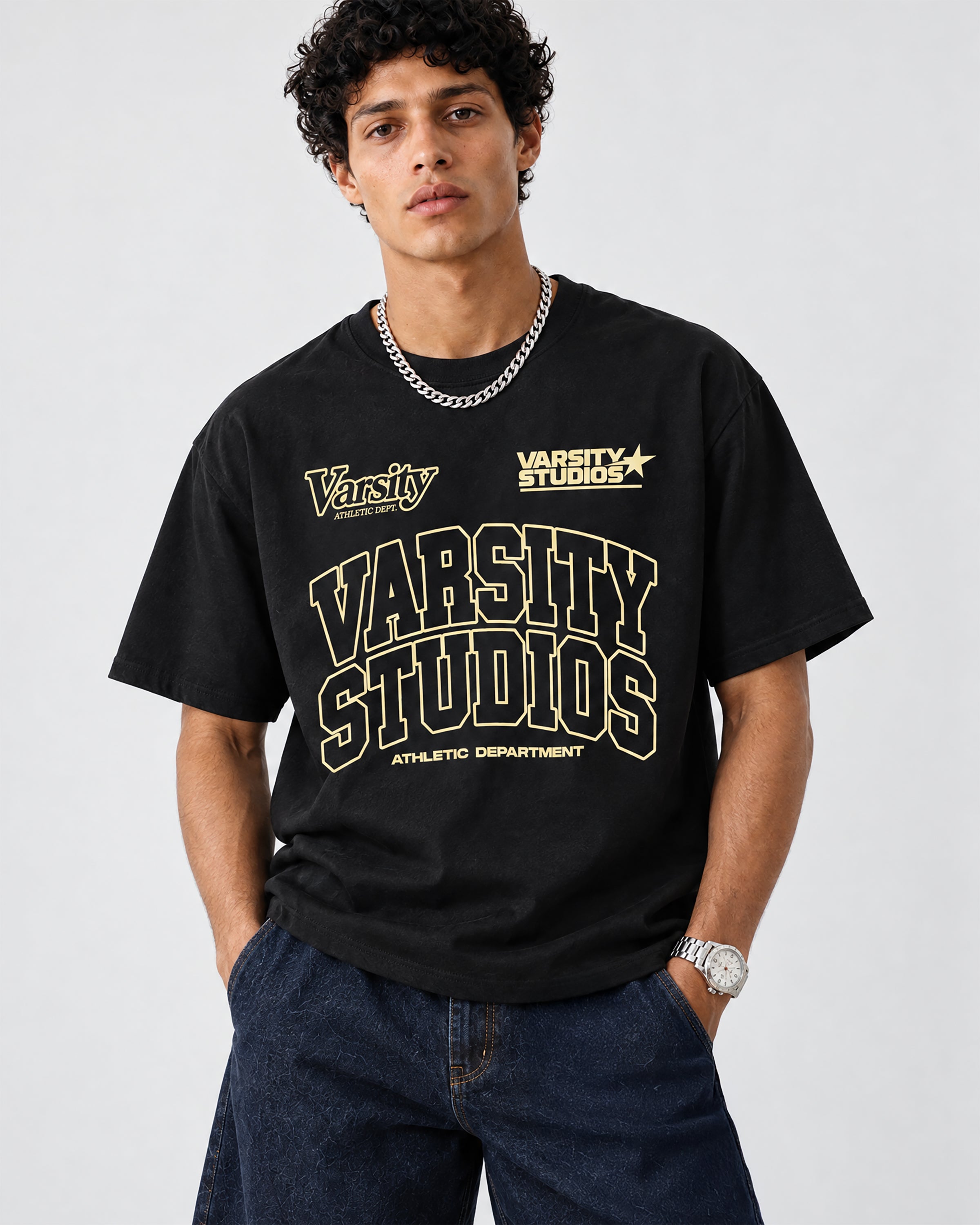 A man wearing a black graphic t-shirt with 'Varsity Studios' printed in gold and white, paired with dark blue jeans, a silver chain necklace, and a silver watch, against a light gray studio background.