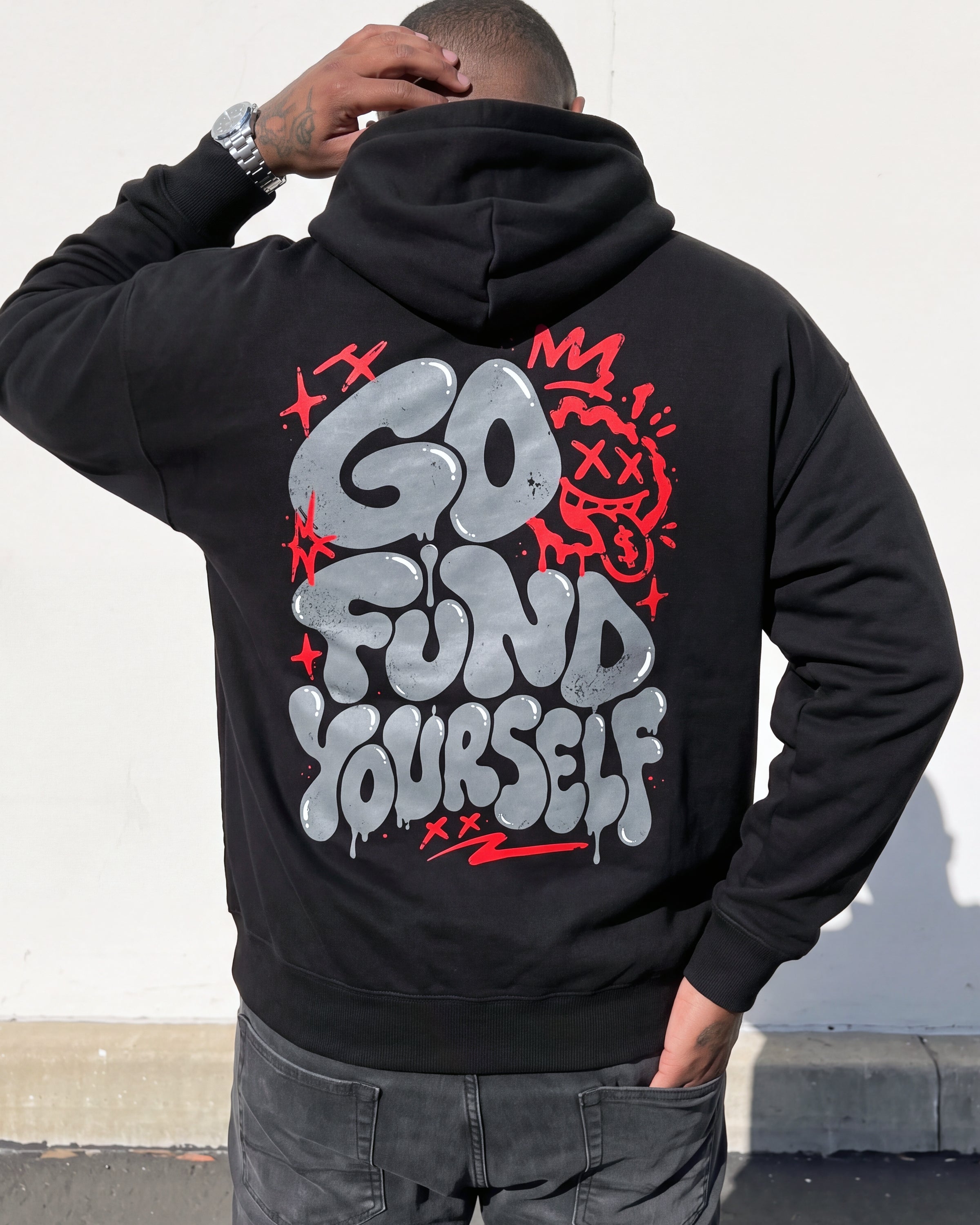 A man viewed from behind wearing a black hoodie with a large graphic on the back that reads 'GO FUND YOURSELF' in silver and red graffiti-style lettering, paired with dark gray jeans.