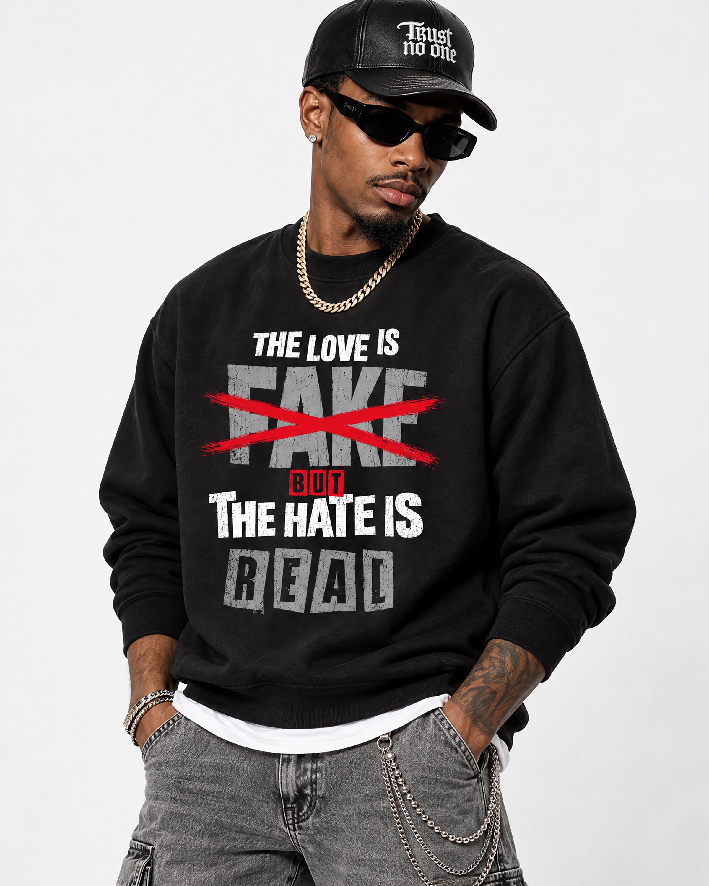 A man wearing a black crewneck sweatshirt with a graphic print that reads 'THE LOVE IS FAKE [crossed out in red] BUT THE HATE IS REAL'. He is also wearing a black cap with 'Trust no one' in white text, black sunglasses, a gold chain necklace, and gray jeans.