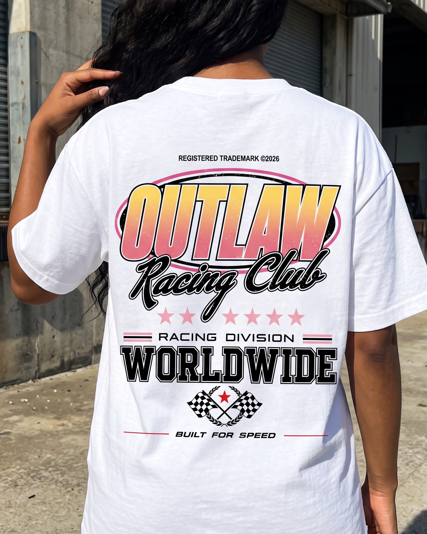 A person viewed from behind wearing a white graphic T-shirt with a large 'Outlaw Racing Club' design in pink, yellow, black, and red on the back, standing outdoors near a concrete wall.