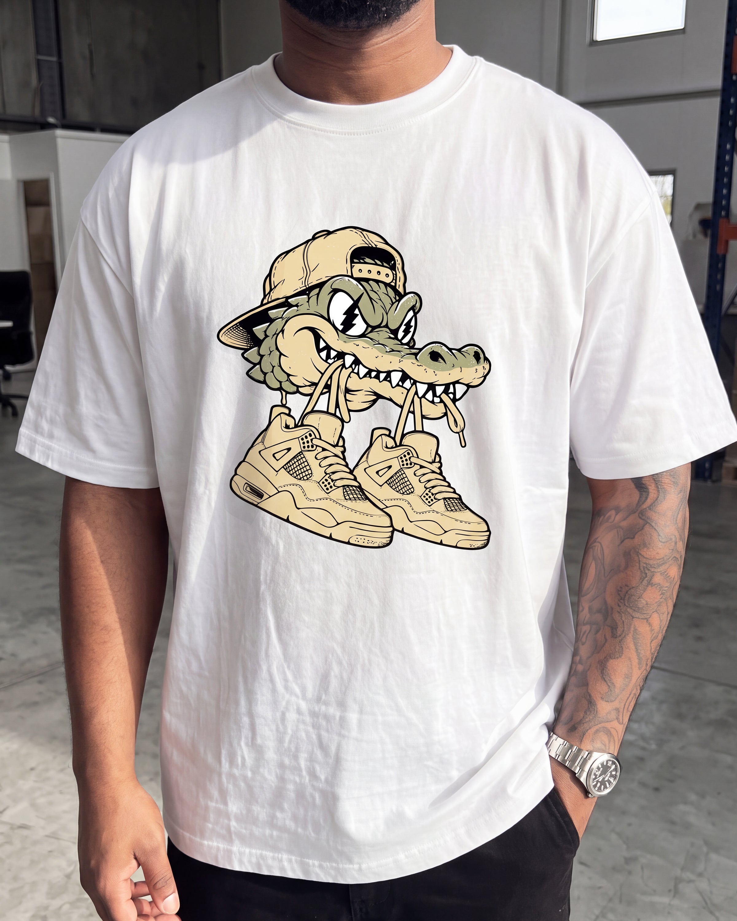 A man wearing a white short-sleeve t-shirt with a cartoon alligator graphic wearing a tan baseball cap and beige sneakers, standing in an indoor warehouse-like setting.