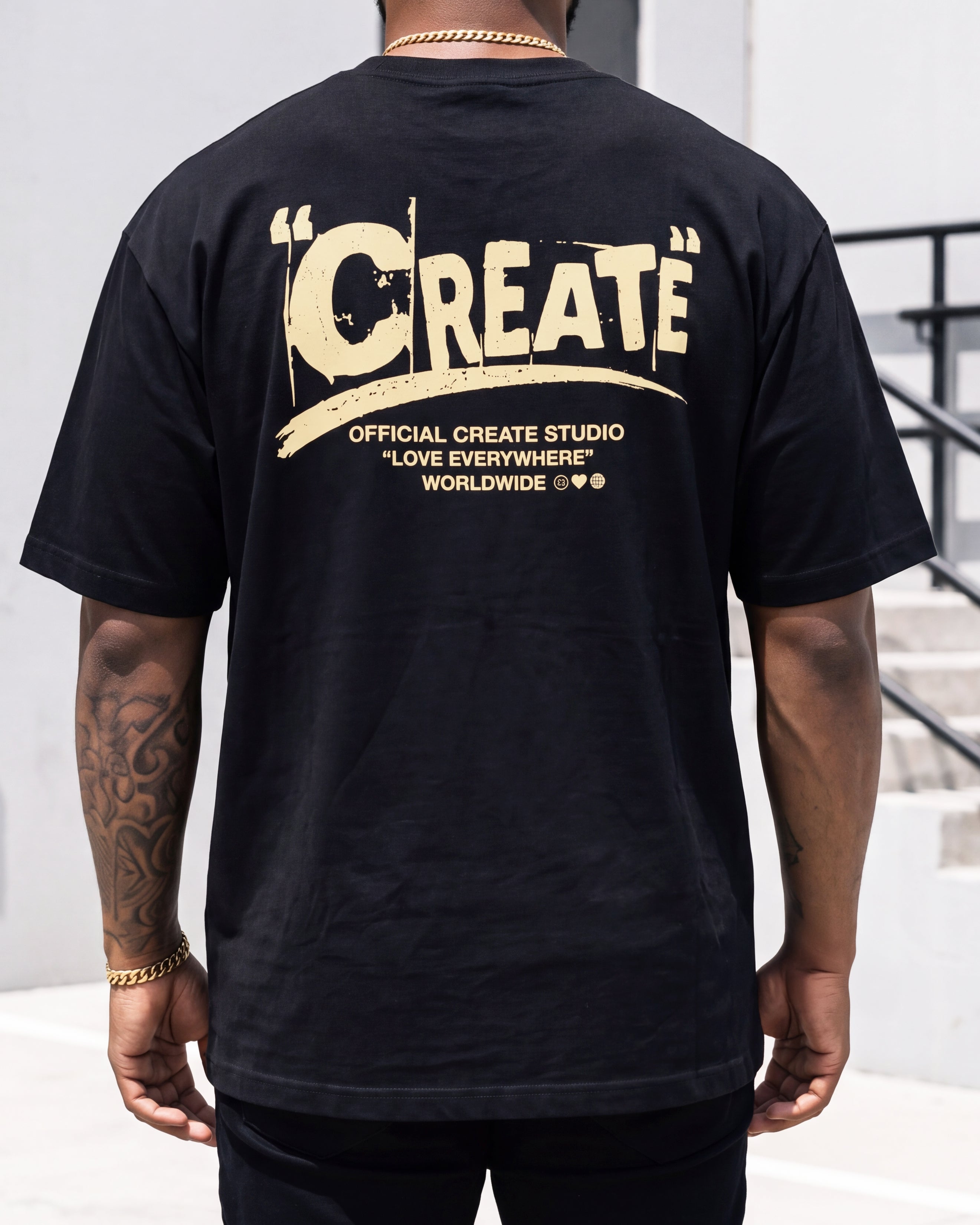 A man viewed from behind wearing a black t-shirt with a large distressed beige graphic on the back that reads 'CREATE' and 'OFFICIAL CREATE STUDIO "LOVE EVERYWHERE" WORLDWIDE'.