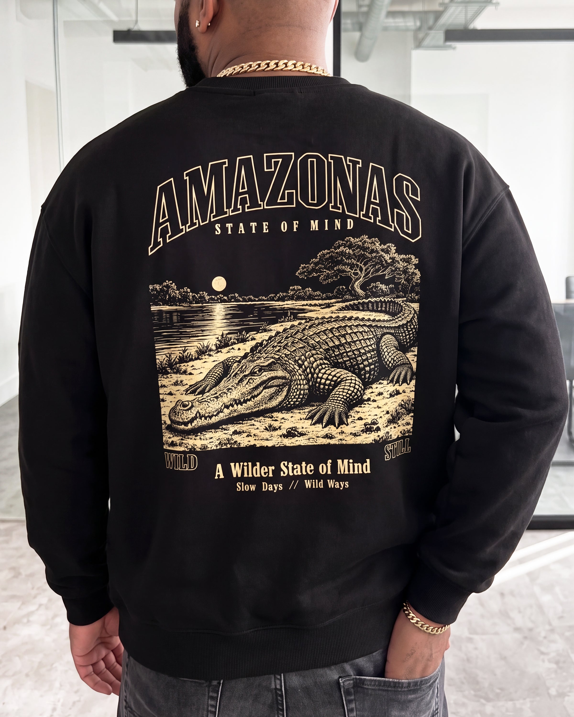 A man viewed from behind wearing a black crewneck sweatshirt with a large tan graphic print on the back featuring an alligator, trees, and the text 'AMAZONAS STATE OF MIND'.