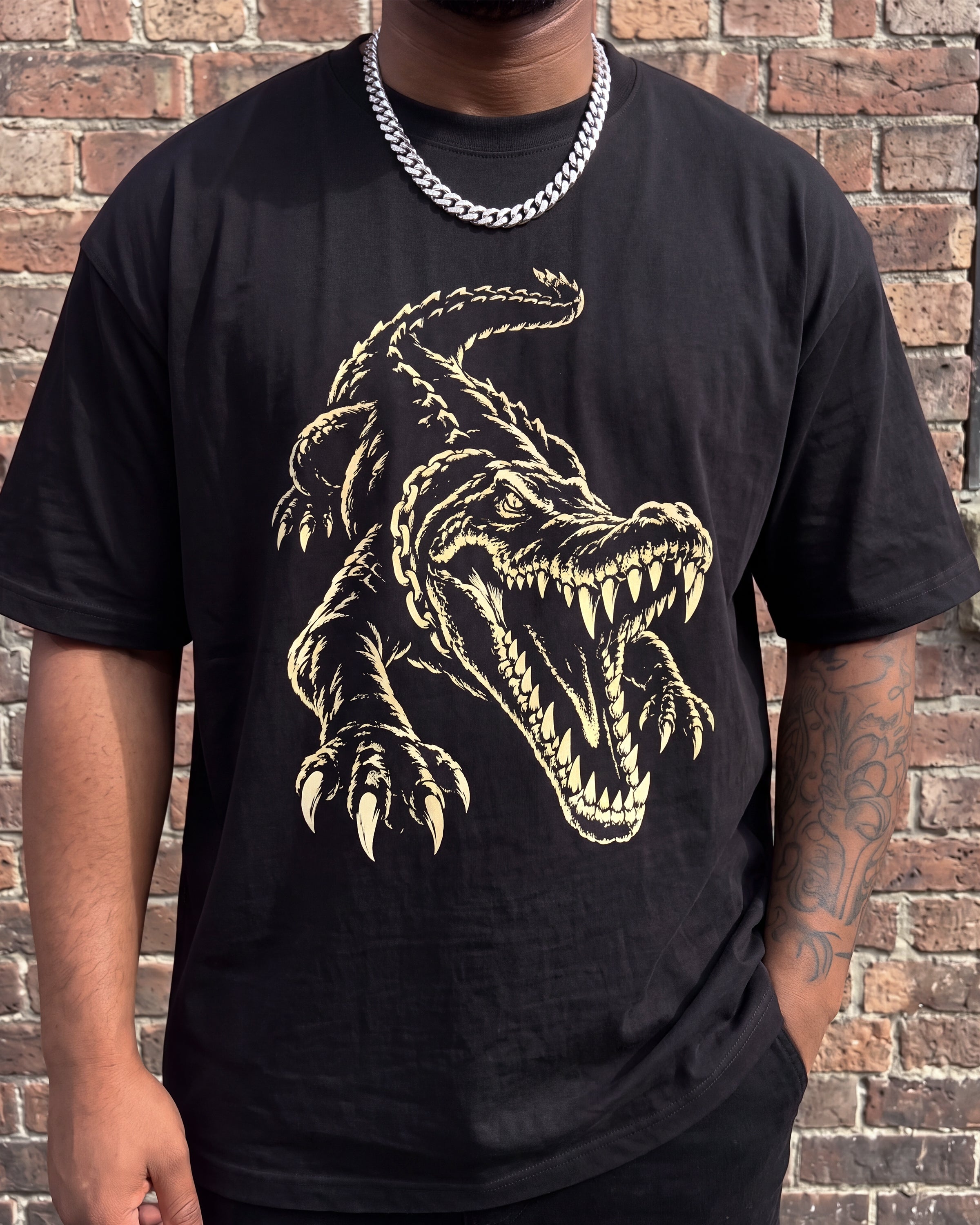 A person wearing a black short-sleeve graphic T-shirt with a large gold crocodile design on the front, standing against a brick wall.