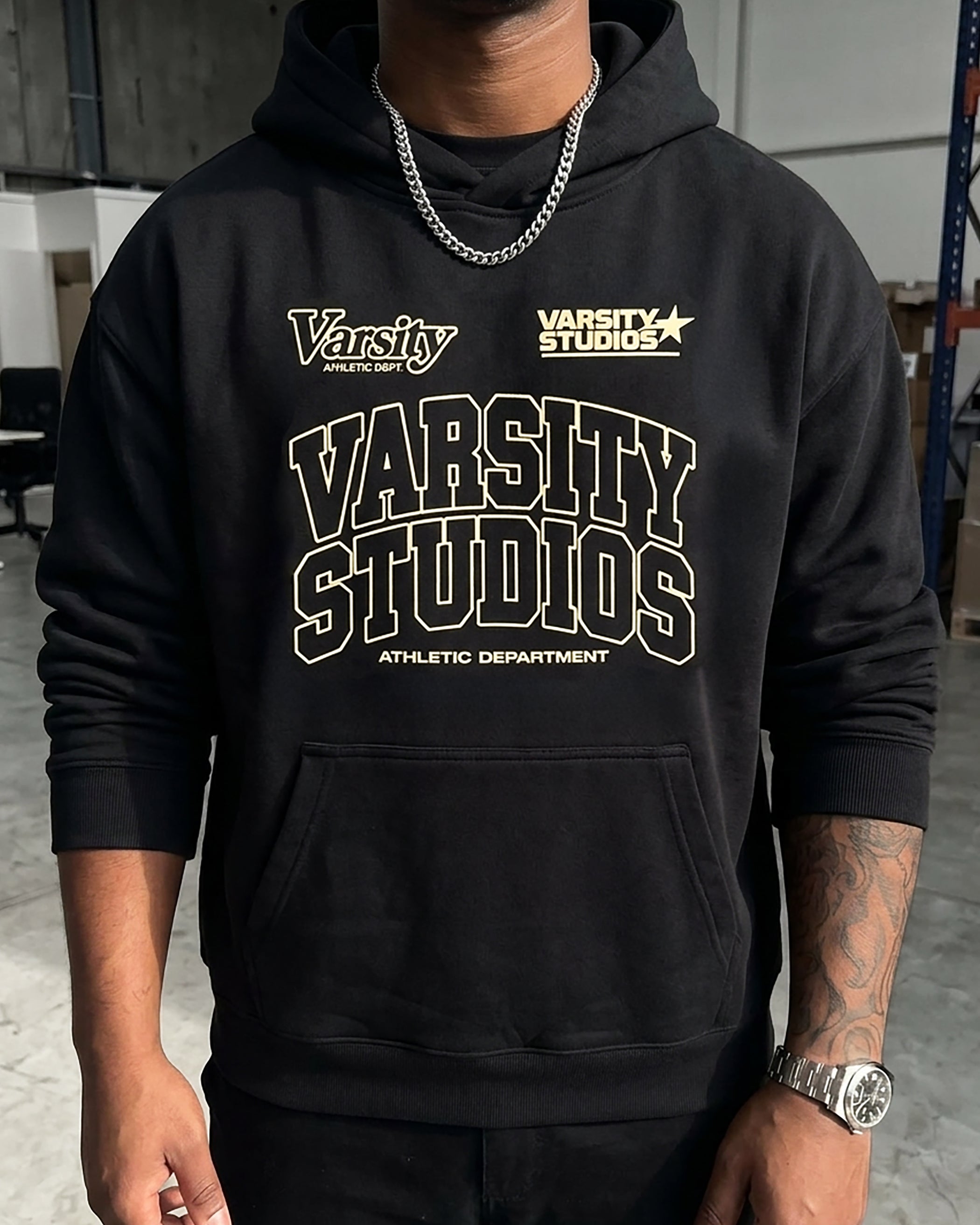 A man wearing a black pullover hoodie with a large 'Varsity Studios Athletic Department' graphic printed in cream and gold on the front, paired with a silver chain necklace.