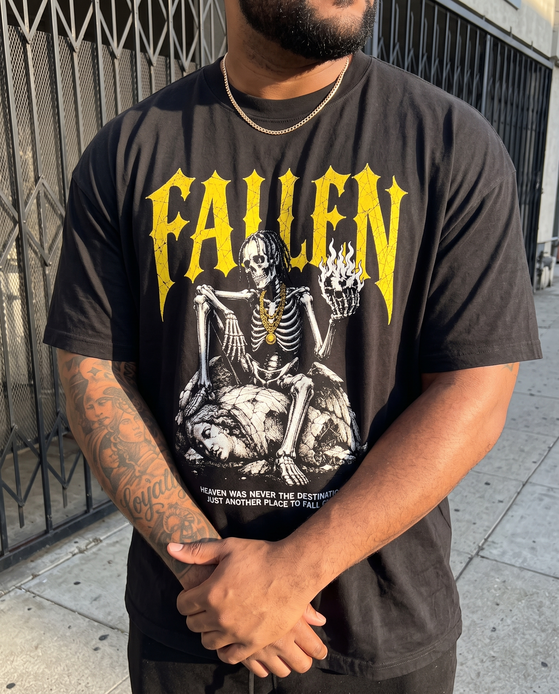 A man wearing a black graphic t-shirt with a yellow 'FALLEN' band logo and a black-and-white skeleton illustration, standing outdoors in front of a metal gate.