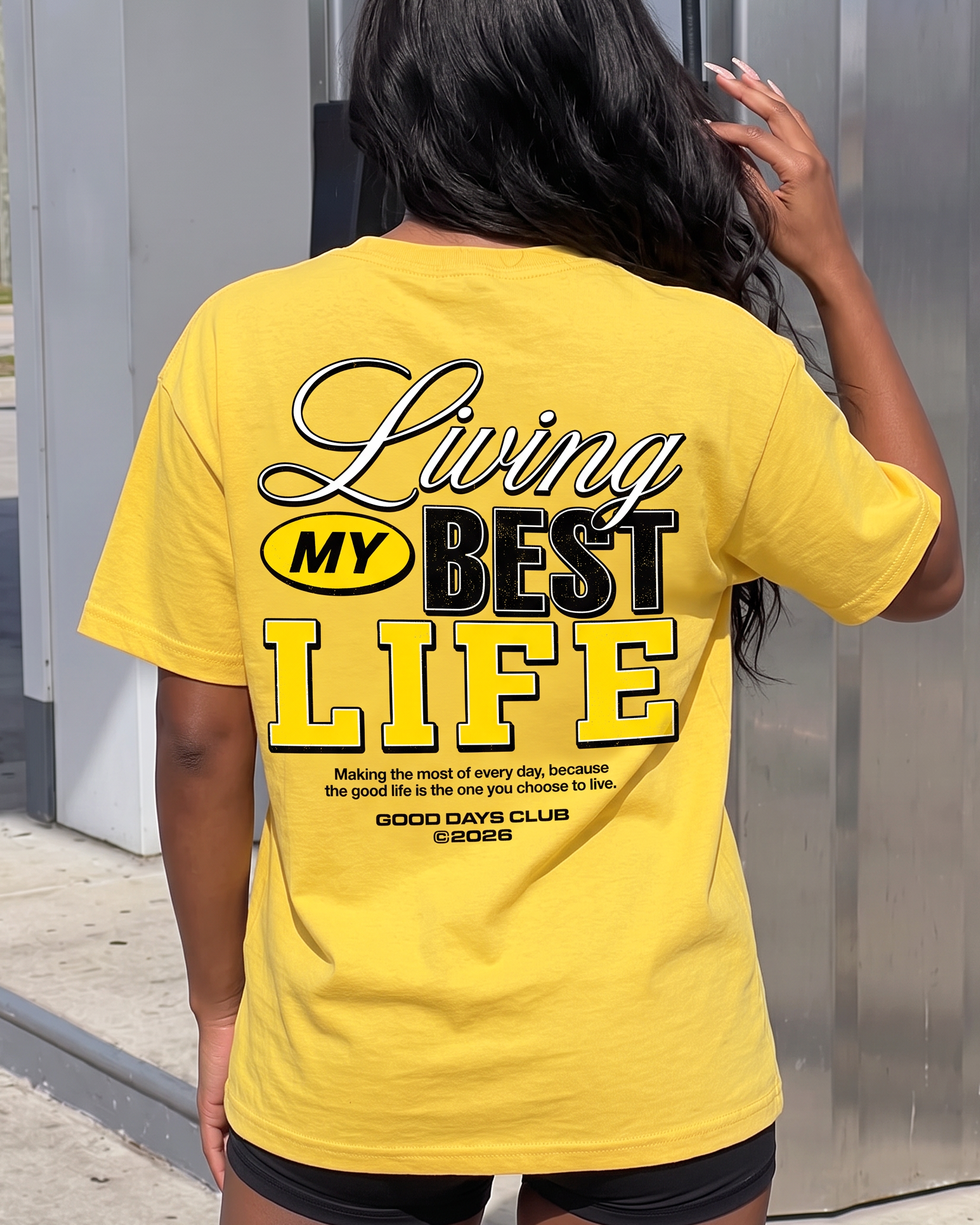 A woman viewed from behind wearing a bright yellow graphic T-shirt with the text 'Living My Best Life' printed on the back, standing outdoors against a gray metal wall.