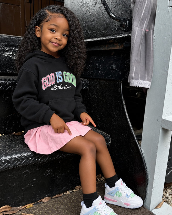 A young girl with curly hair sits on black metal steps, wearing a black hoodie with the text 'GOD IS GOD all the time' in pink, green, and white, paired with a pink pleated skirt and pastel sneakers.