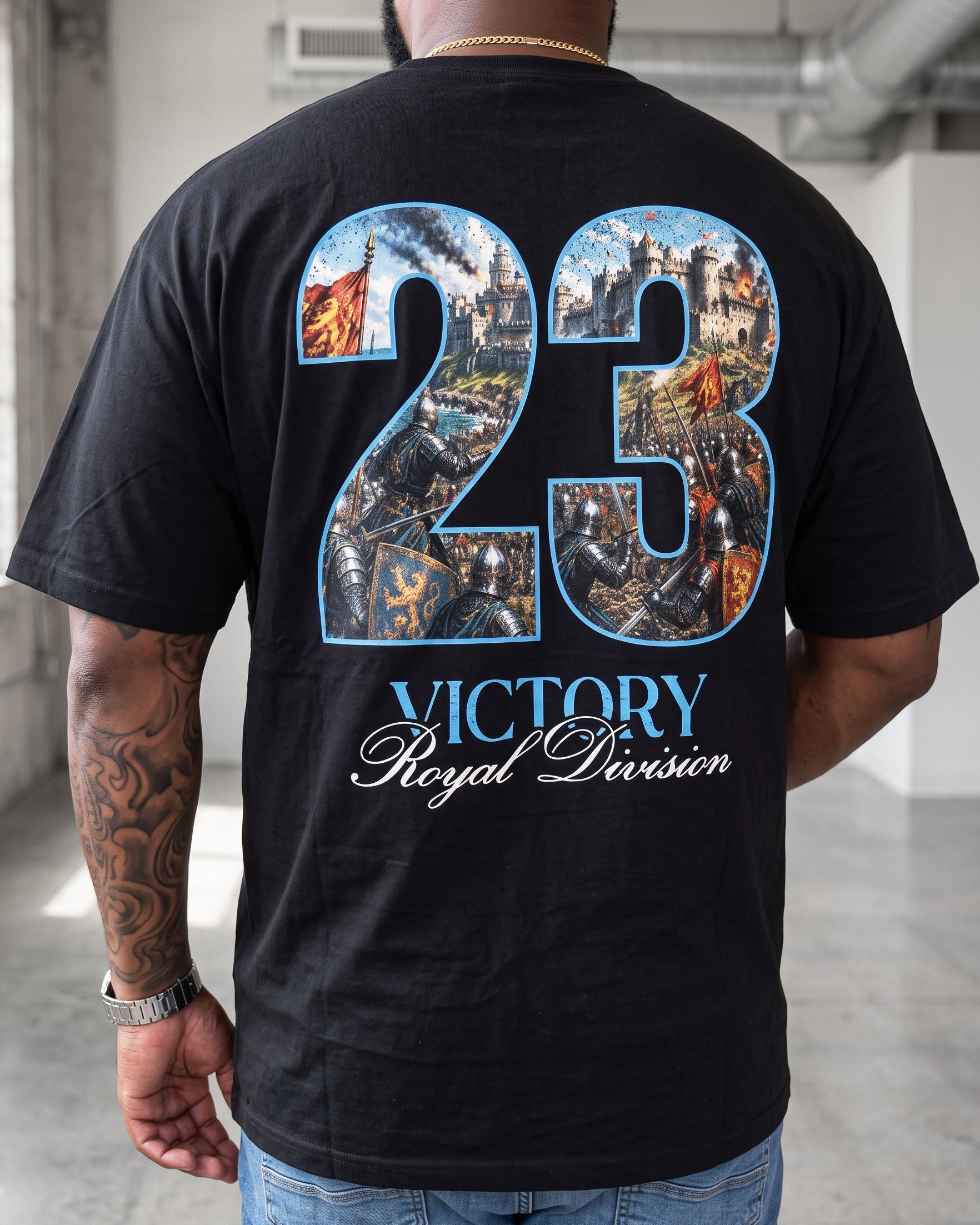 A man viewed from behind wearing a black t-shirt with a large graphic on the back. The graphic features the number '23' filled with a detailed battle scene, outlined in blue, with 'VICTORY Royal Division' printed below in blue and white script.