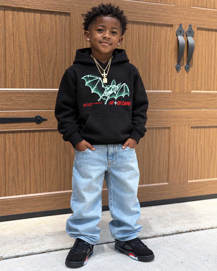 A young boy stands in front of a wooden garage door wearing a black hoodie with a green bat graphic and red 'AIR DARK' text, light blue jeans, and black sneakers.