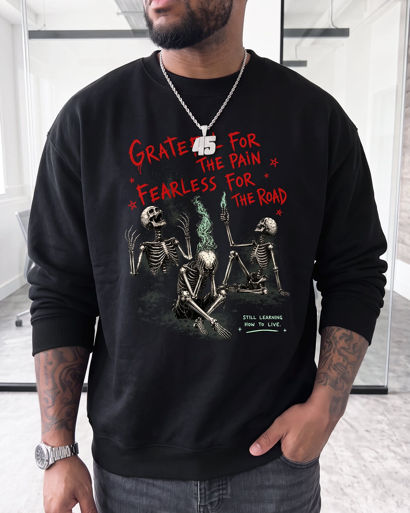 A man wearing a black crewneck sweatshirt with a graphic print of three skeletons and the text 'GRATE FOR THE PAIN FEARLESS FOR THE ROAD' in red, paired with gray jeans and a silver chain necklace.