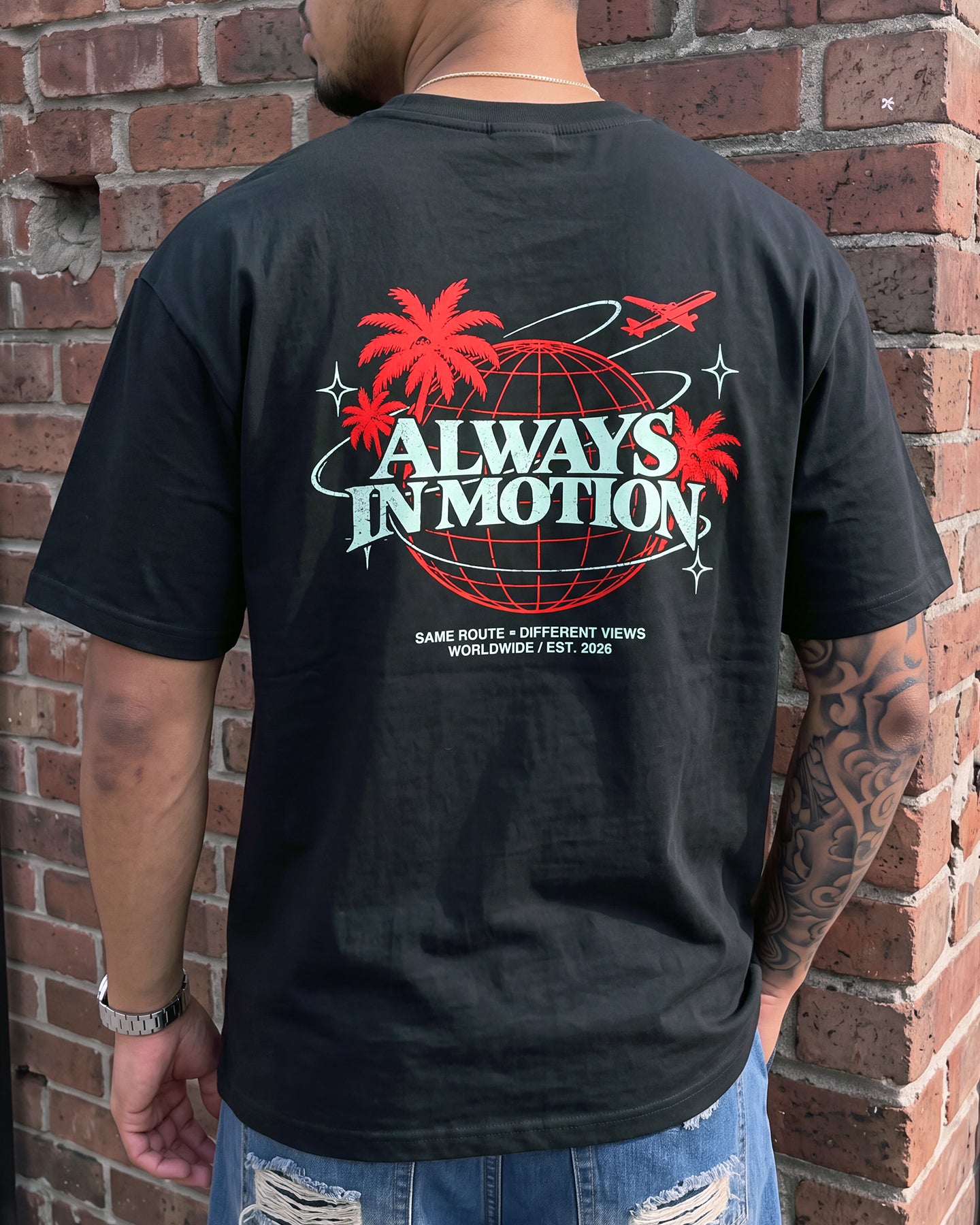 A man viewed from behind wearing a black graphic t-shirt with a large retro design on the back featuring the text 'ALWAYS IN MOTION' in white and red, with palm trees and a globe. He is standing against a red brick wall.