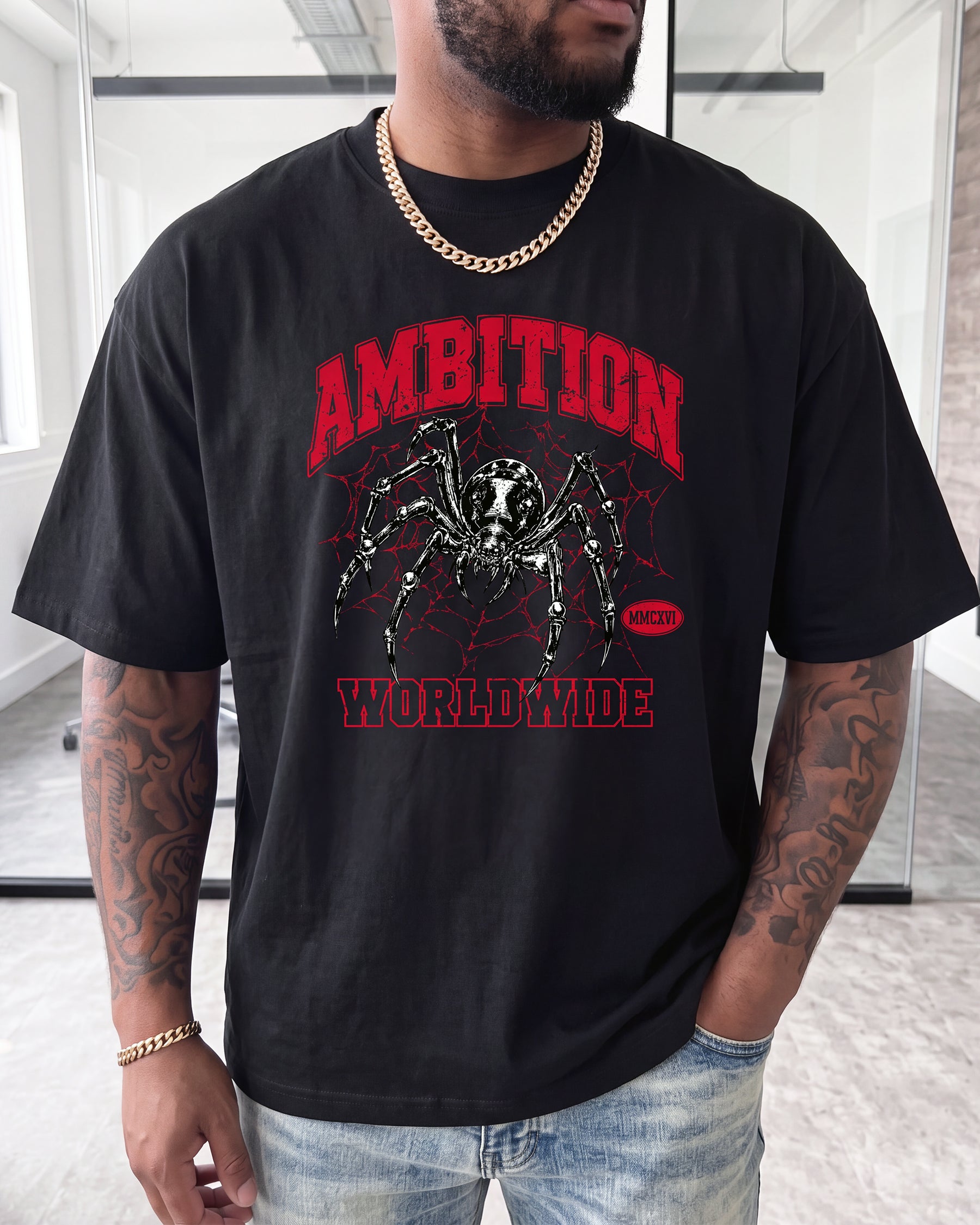 A man wearing a black graphic t-shirt with a red and black spider design and the words 'AMBITION' and 'WORLDWIDE' on the front, paired with light blue jeans and gold chain jewelry.
