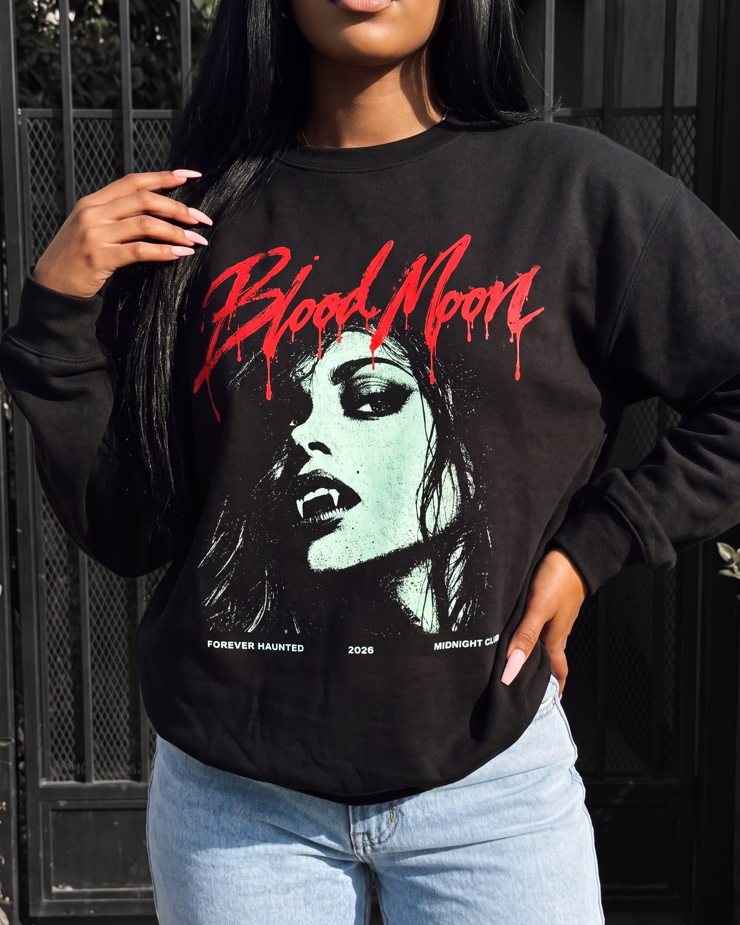 A woman wearing a black crewneck sweatshirt with a graphic print of a vampire woman's face and the words 'Blood Moon' in red dripping lettering, paired with light blue jeans.