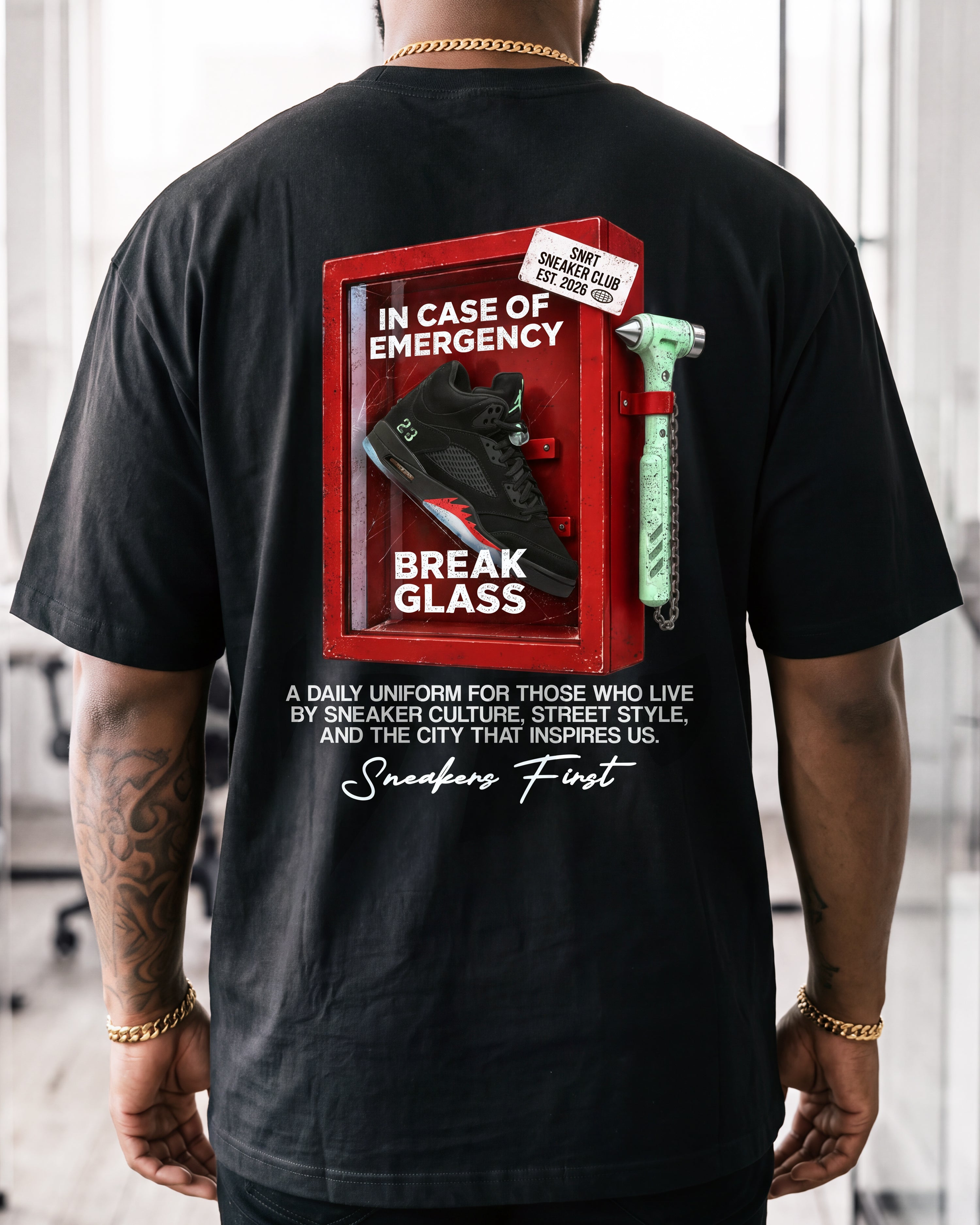 A man wearing a black t-shirt from the back, featuring a graphic print of a red emergency break glass box with a sneaker and a green chain, with the text 'Sneakers First'.
