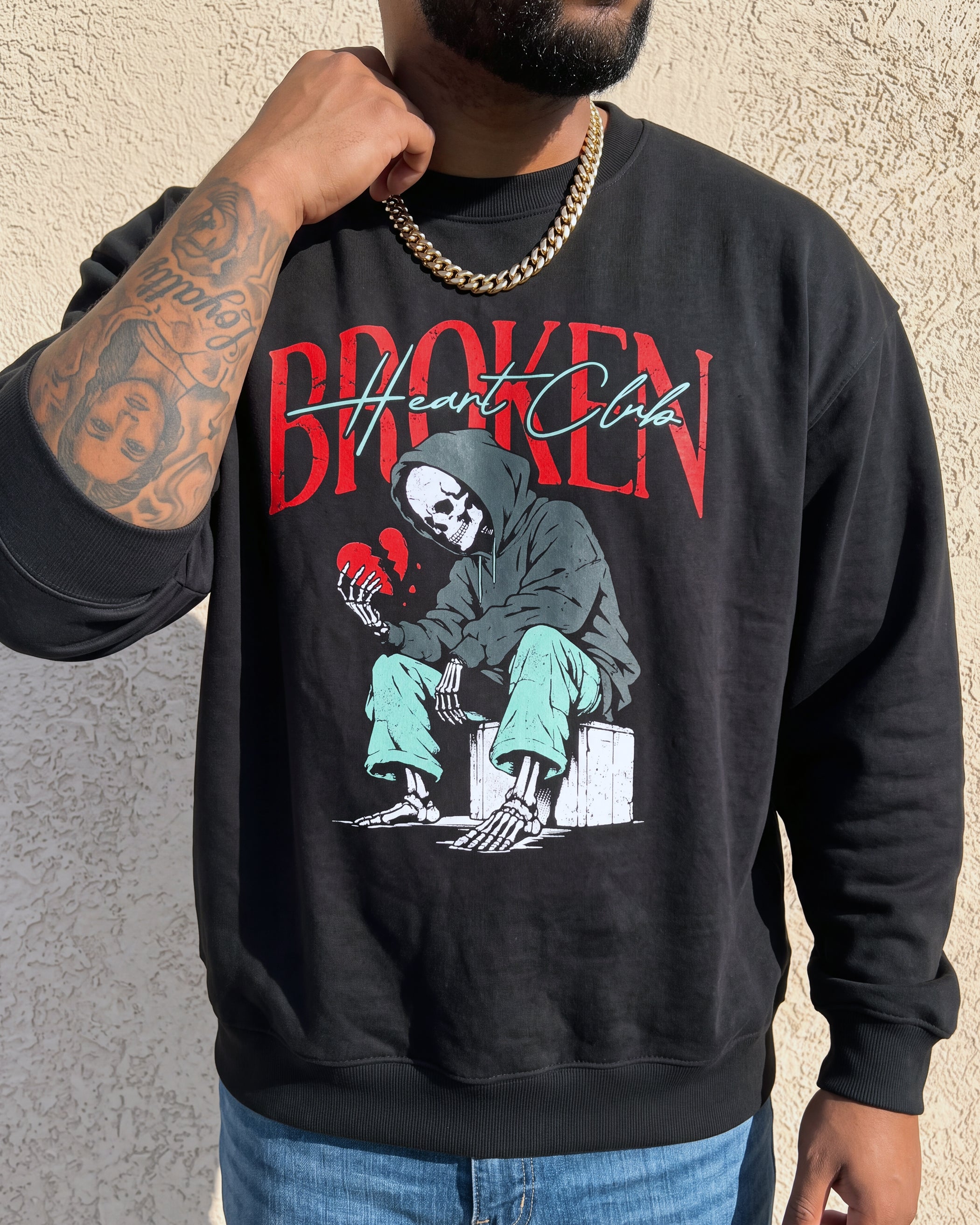 A man wearing a black crewneck sweatshirt with a graphic print of a skeleton holding a broken red heart and the text 'Broken Heart Club'.