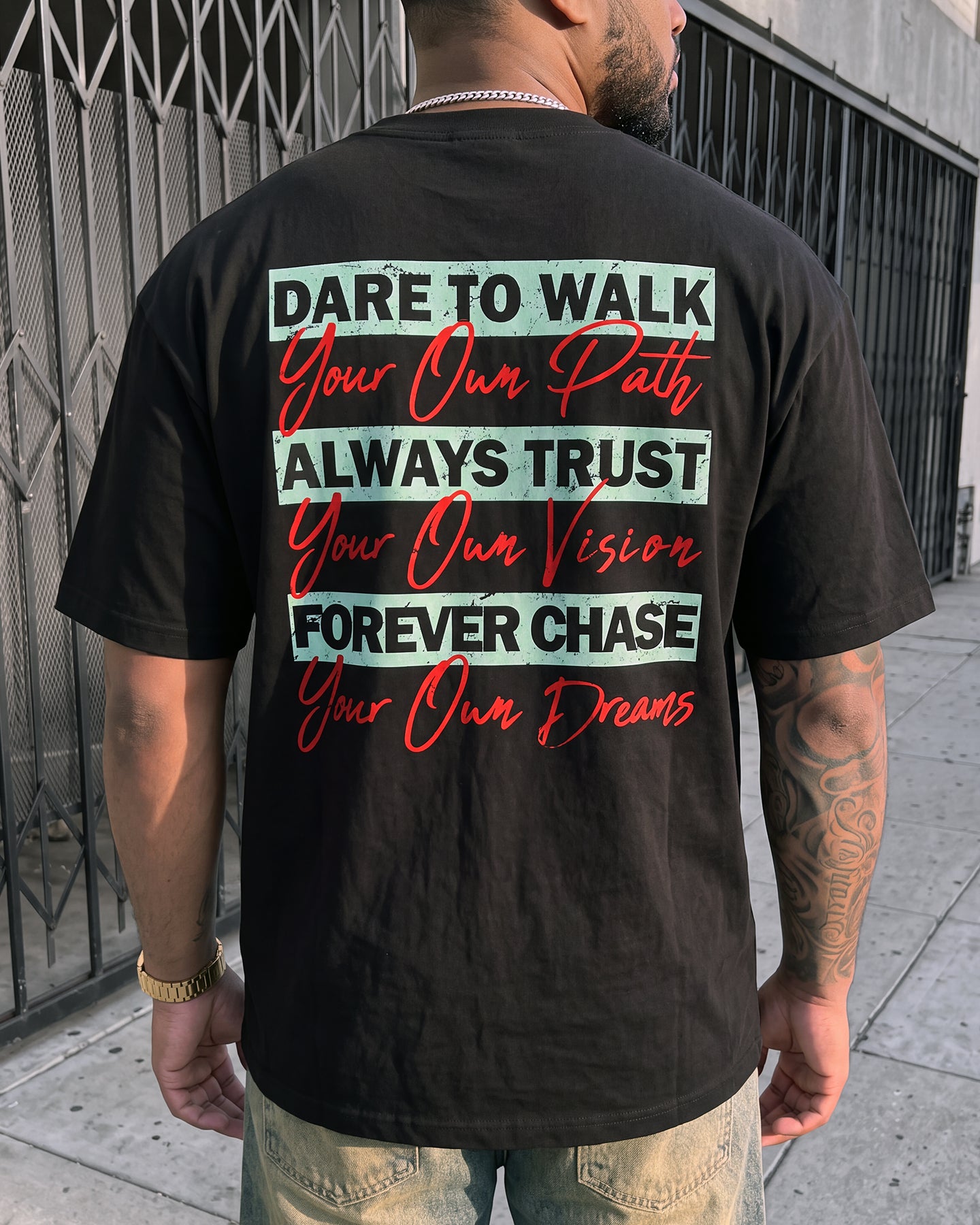 A man viewed from behind wearing a black graphic t-shirt with motivational text on the back, standing on a sidewalk in front of a metal gate.
