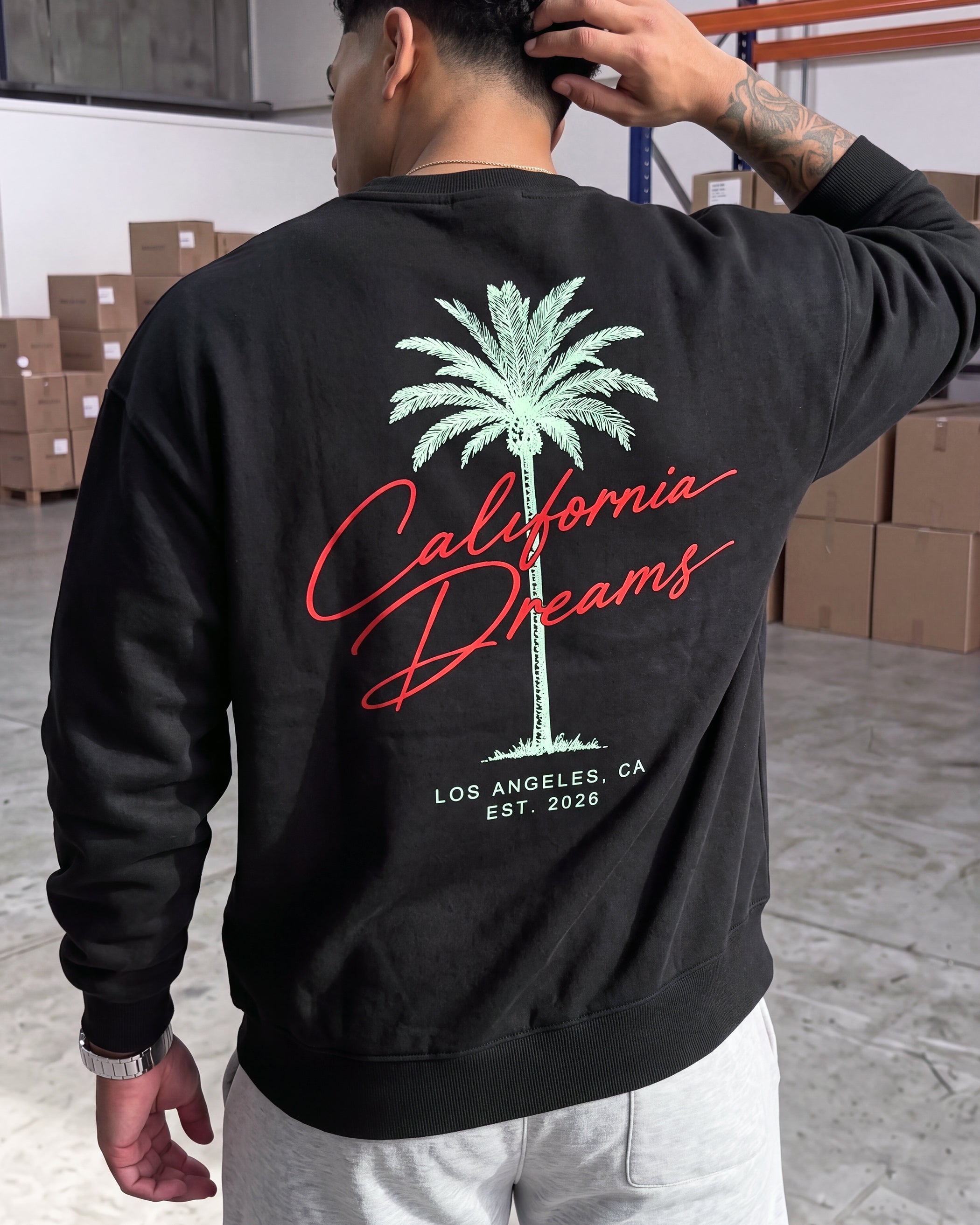 A man wearing a black crewneck sweatshirt with a graphic on the back. The graphic features a green palm tree, red cursive text reading 'California Dreams', and white text reading 'LOS ANGELES, CA EST.