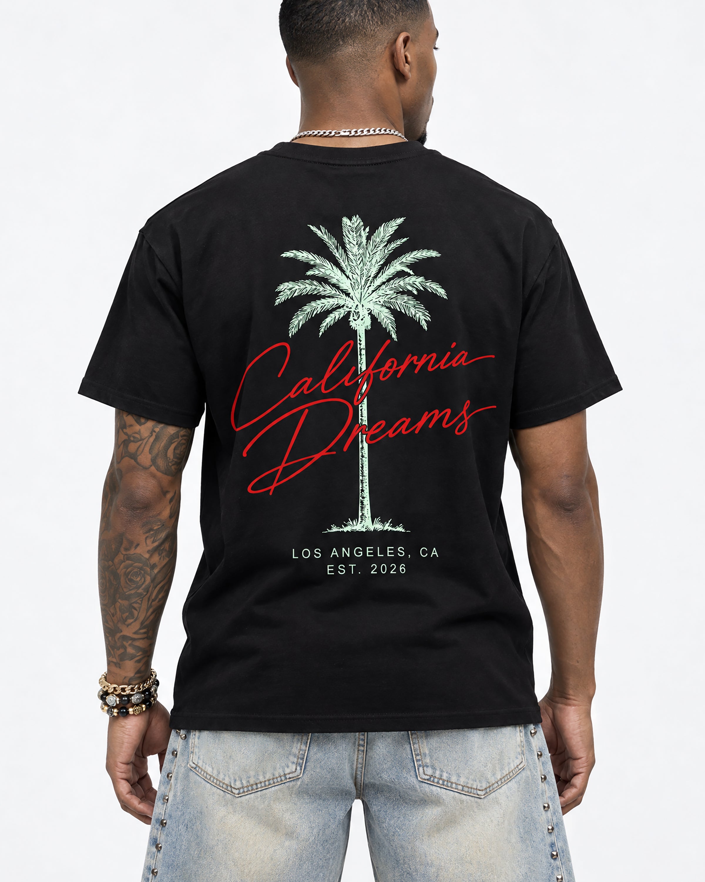A man viewed from the back wearing a black t-shirt with a large graphic print of a palm tree, red cursive text reading 'California Dreams', and white text reading 'Los Angeles, CA Est. 2026'.