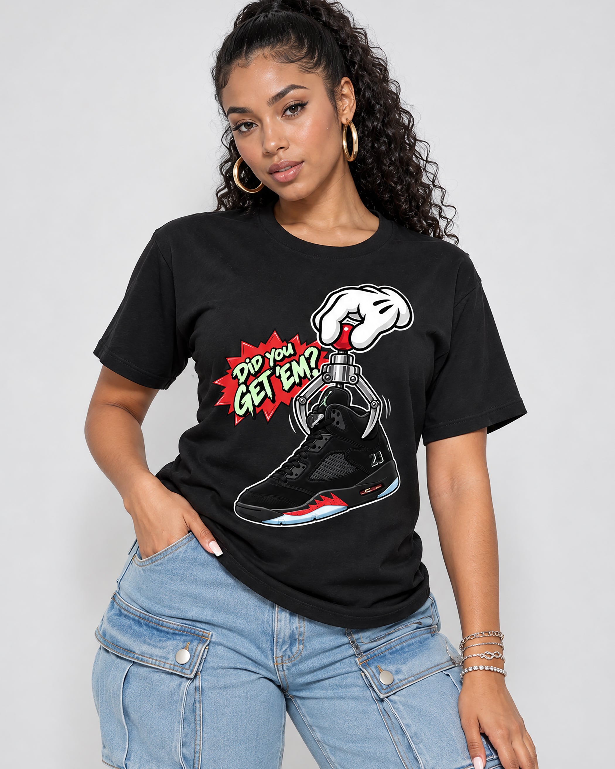 A woman wearing a black graphic t-shirt with a comic-style design that reads 'Did You GET EMP?' and shows a sneaker being crushed by a fist. She is paired with light blue jeans against a plain light gray background.