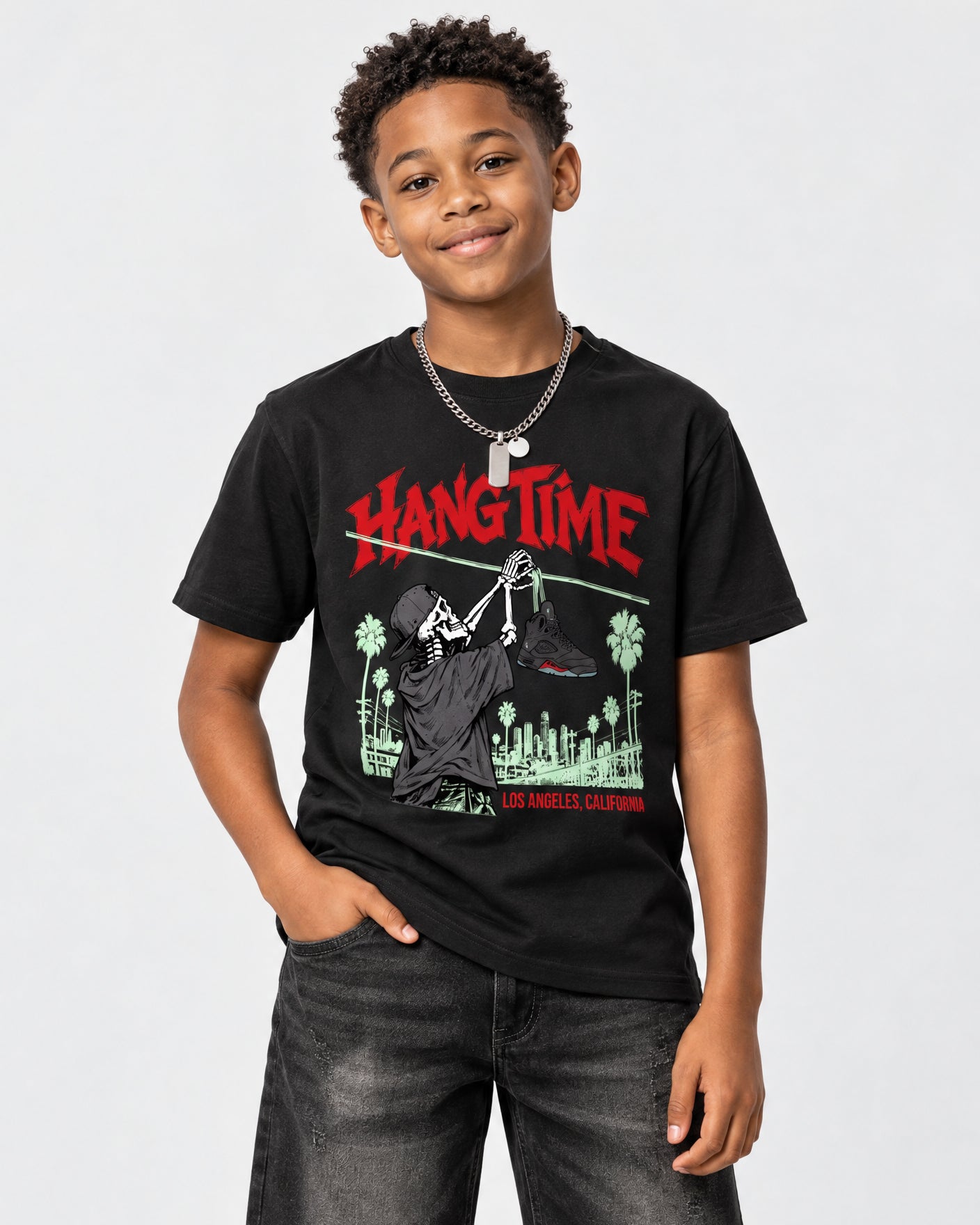 A young boy wearing a black graphic t-shirt with a red and green 'Hang Time' design, paired with dark gray jeans, standing against a light gray studio background.