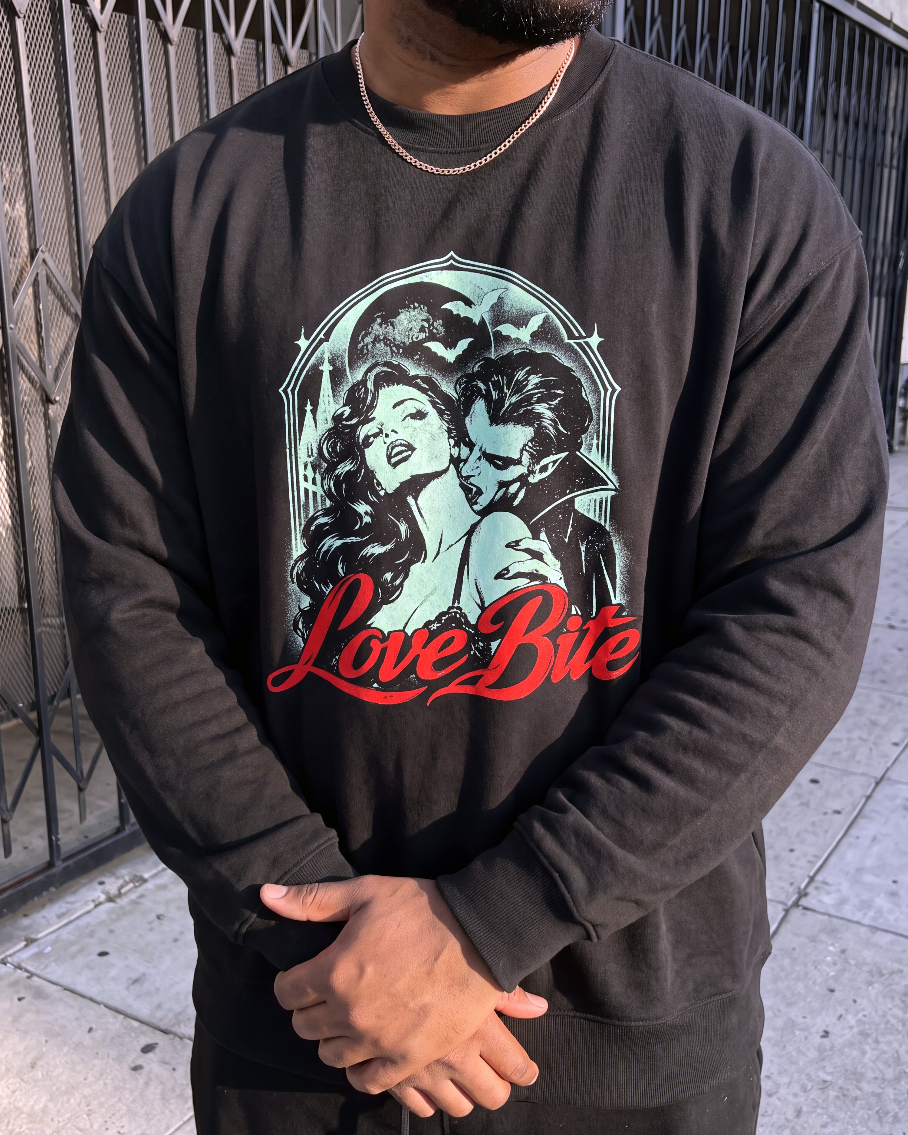 A person wearing a black crewneck sweatshirt with a vintage-style graphic of a woman and man with the text 'Love Bite' in red, standing outdoors near a metal gate.