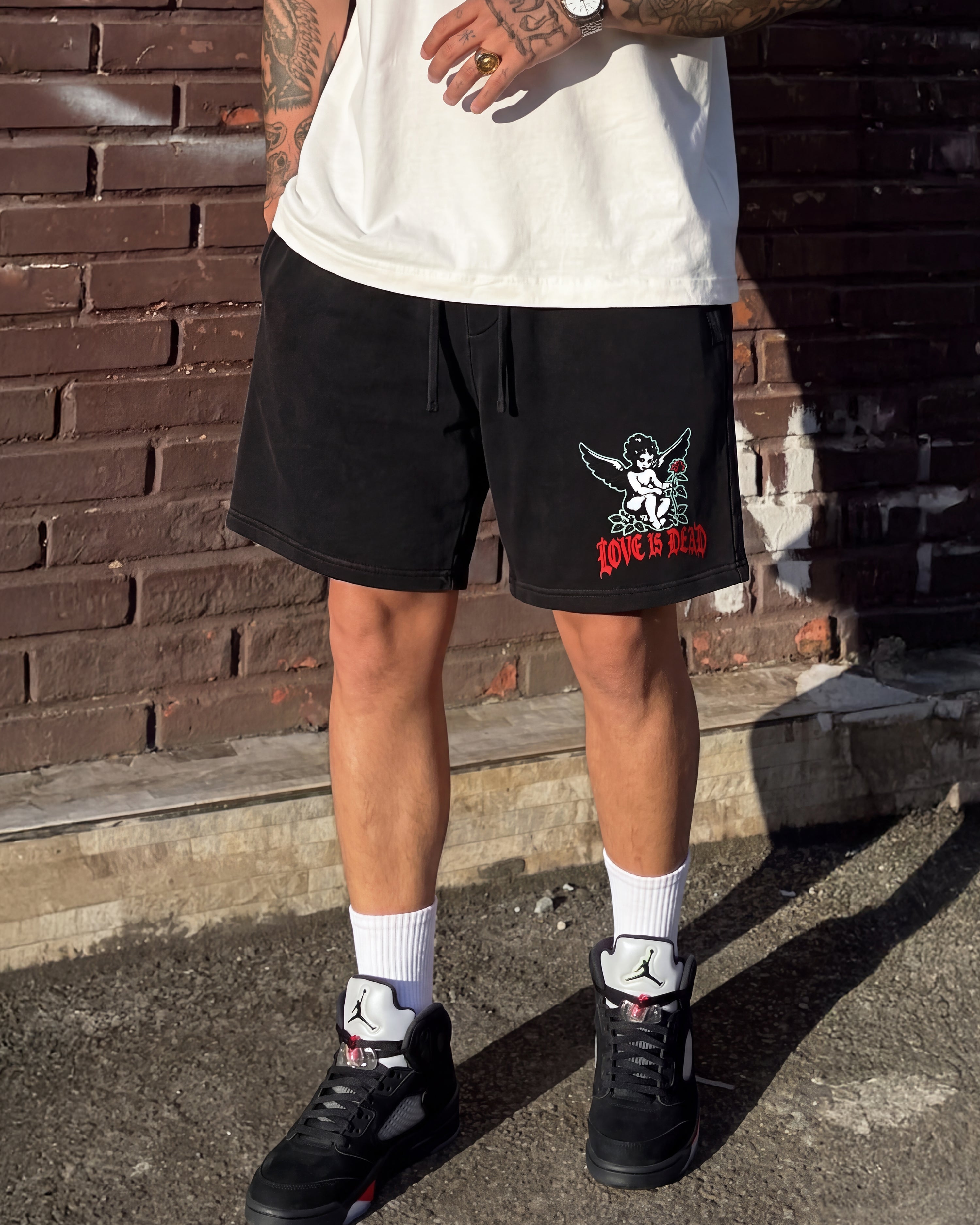A person wearing black drawstring shorts with a red and white graphic on the left leg, paired with a white t-shirt, white socks, and black sneakers, standing outdoors against a brick wall.