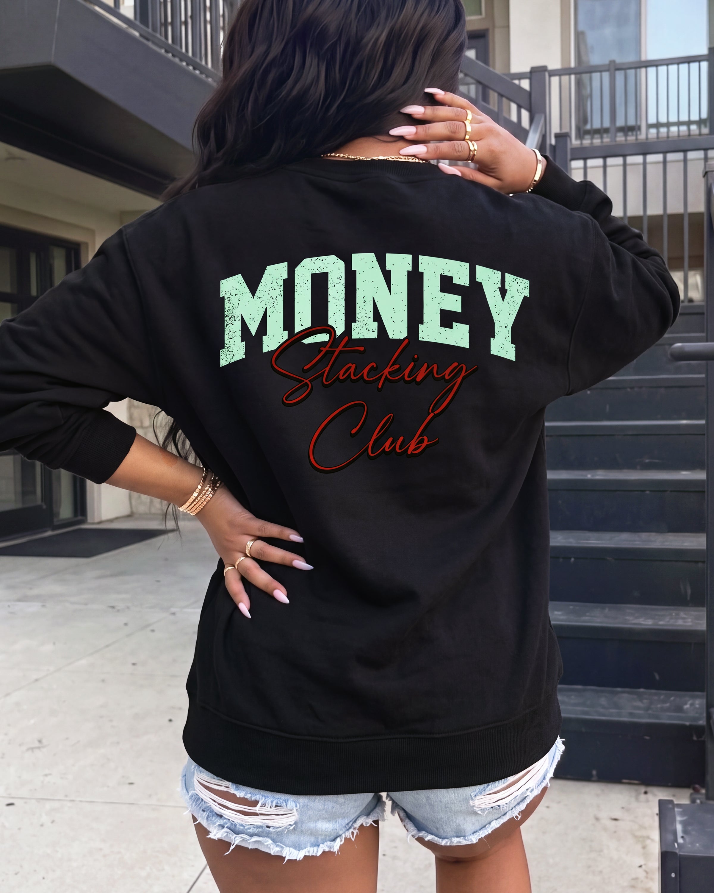 A woman viewed from behind wearing a black sweatshirt with 'MONEY Stacking Club' printed on the back in mint green and red, paired with distressed denim shorts, standing outdoors in front of a building with stairs.