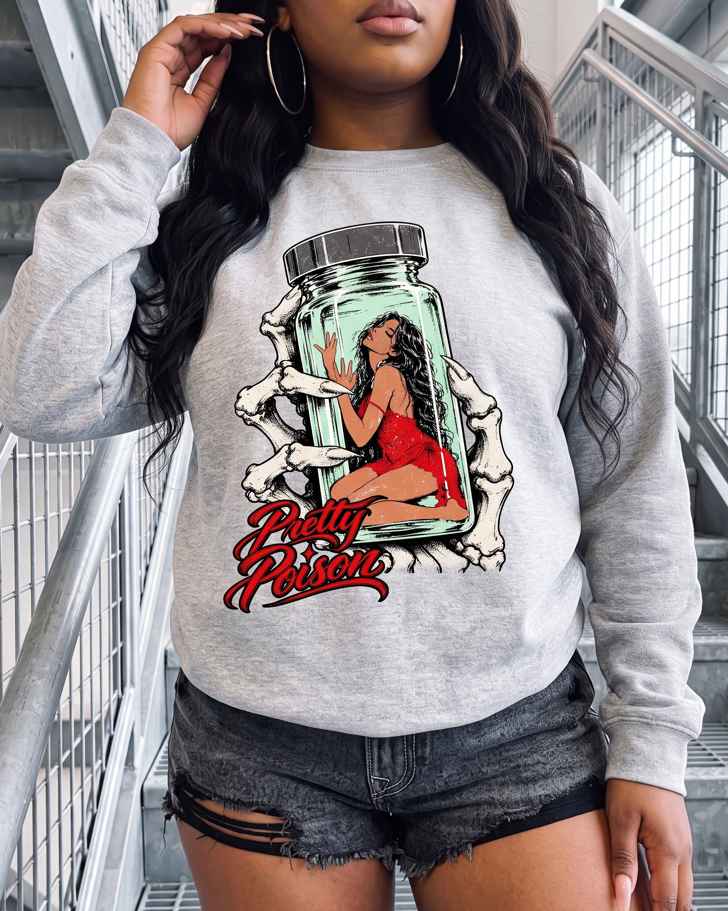 A woman wearing a light gray heather crewneck sweatshirt with a graphic print of a woman in a red dress inside a glass jar, surrounded by skeleton hands and the text 'Pretty Poison'. She is also wearing distressed denim shorts.