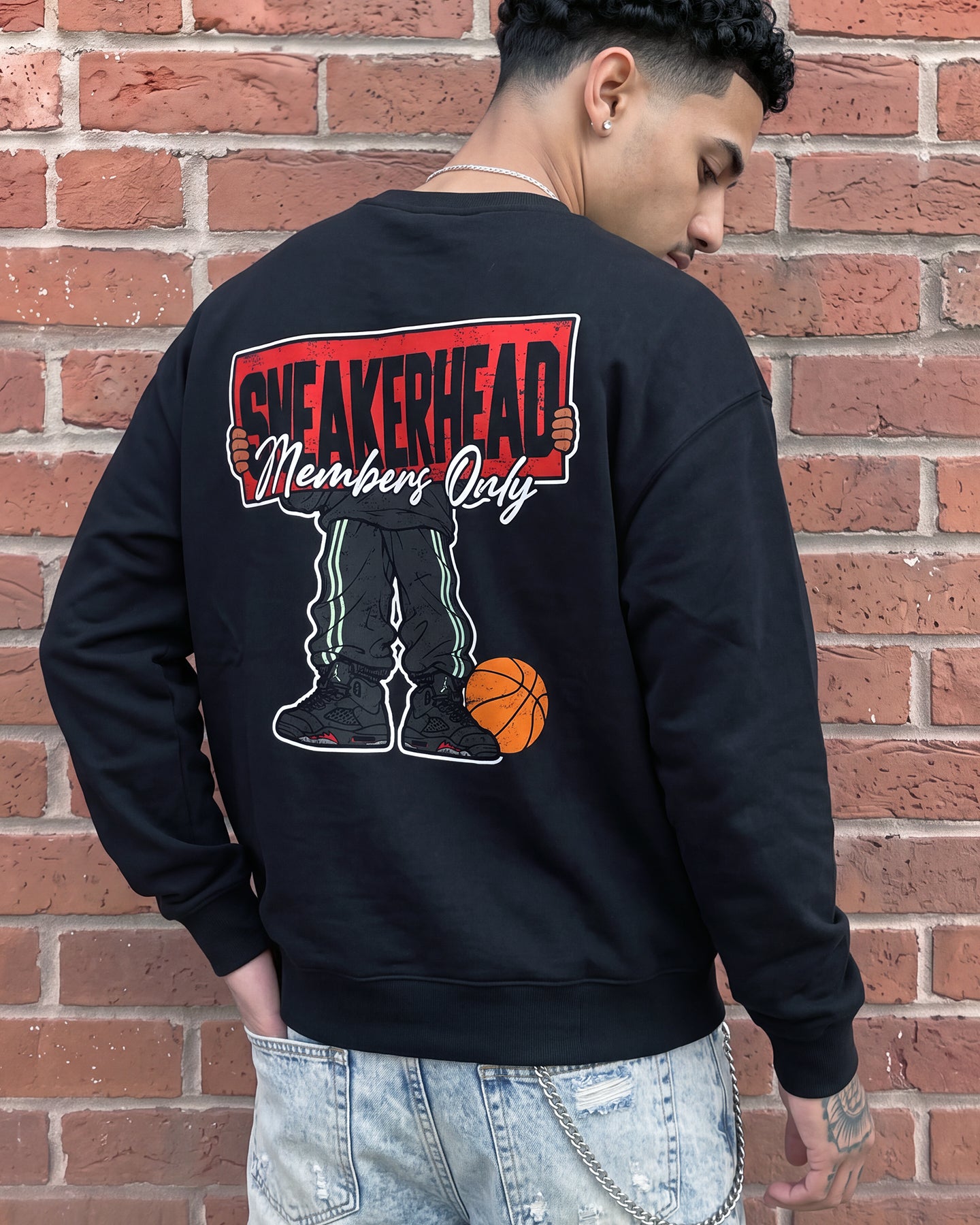 A man wearing a black crewneck sweatshirt with a large back graphic that reads 'SNEAKERHEAD Members Only' above a cartoon figure with green pants and an orange basketball, standing against a red brick wall.