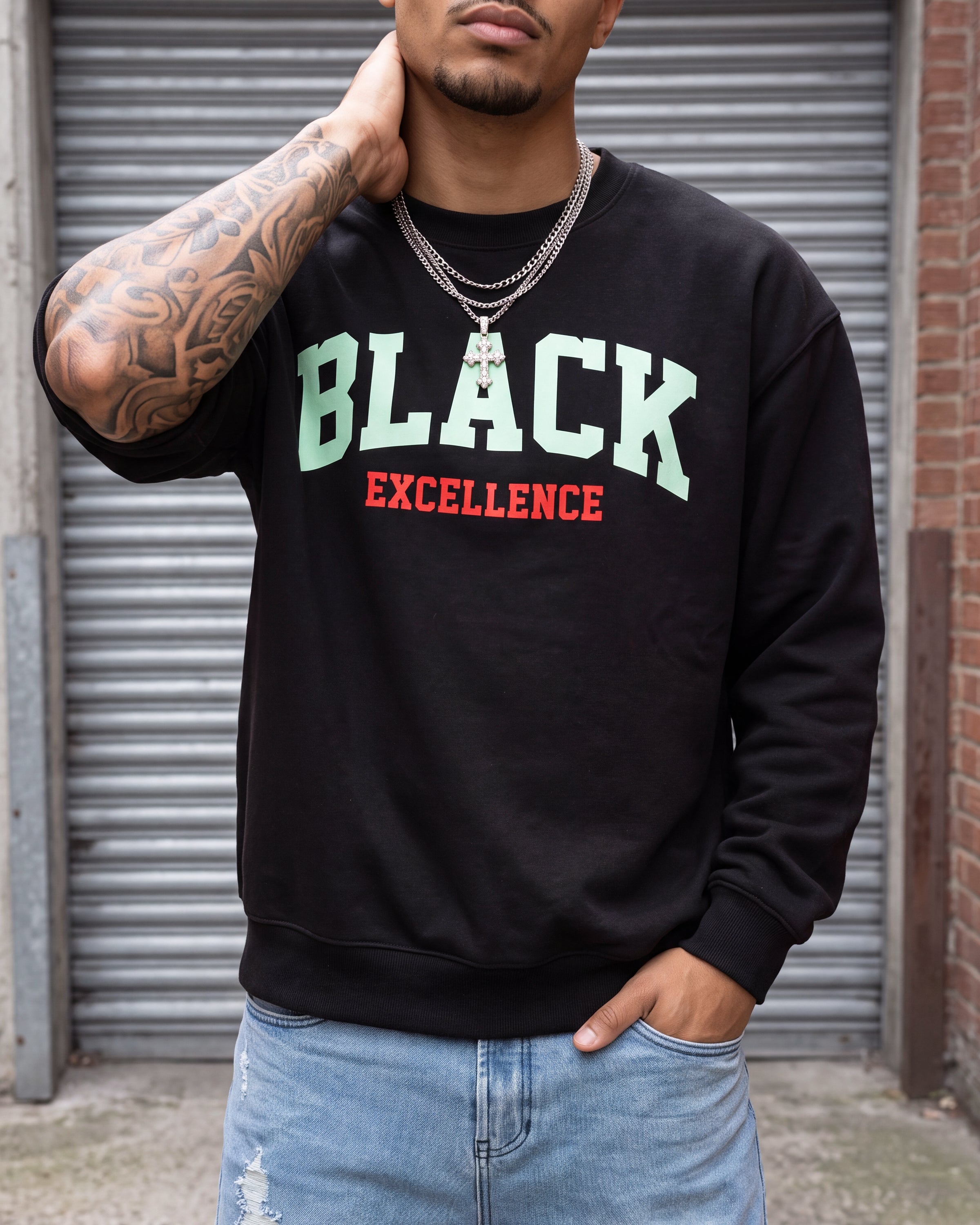 A man wearing a black crewneck sweatshirt with 'BLACK EXCELLENCE' printed on the chest in light green and red, paired with light blue jeans, standing in front of a gray metal garage door.
