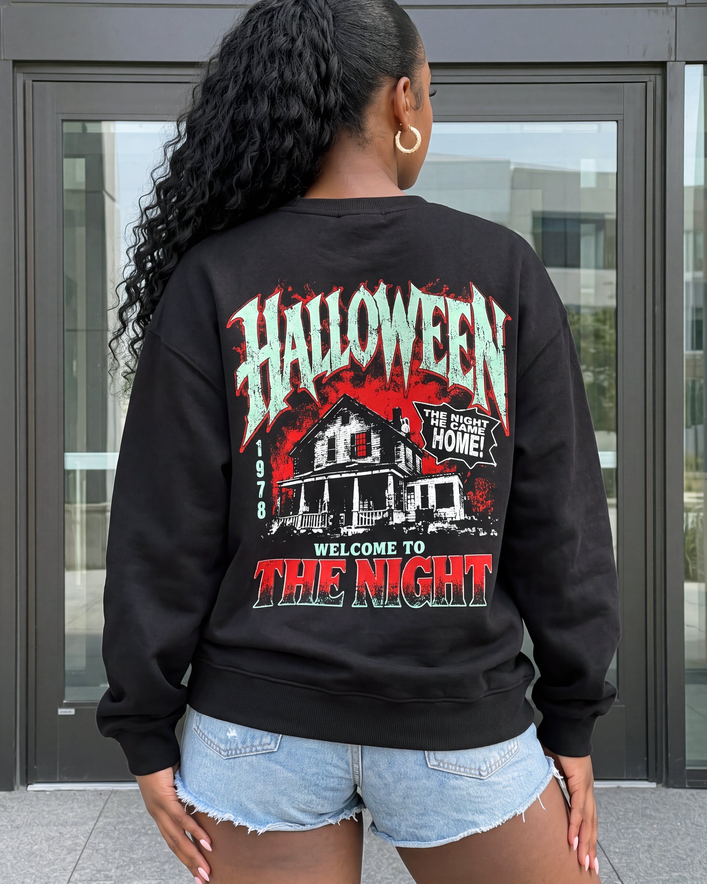 A woman viewed from behind wearing a black crewneck sweatshirt with a large Halloween-themed graphic on the back. The graphic features the word 'HALLOWEEN' in a distressed white and red font above an illustration of a haunted house, with the text 'WELCOME TO THE NIGHT' below.