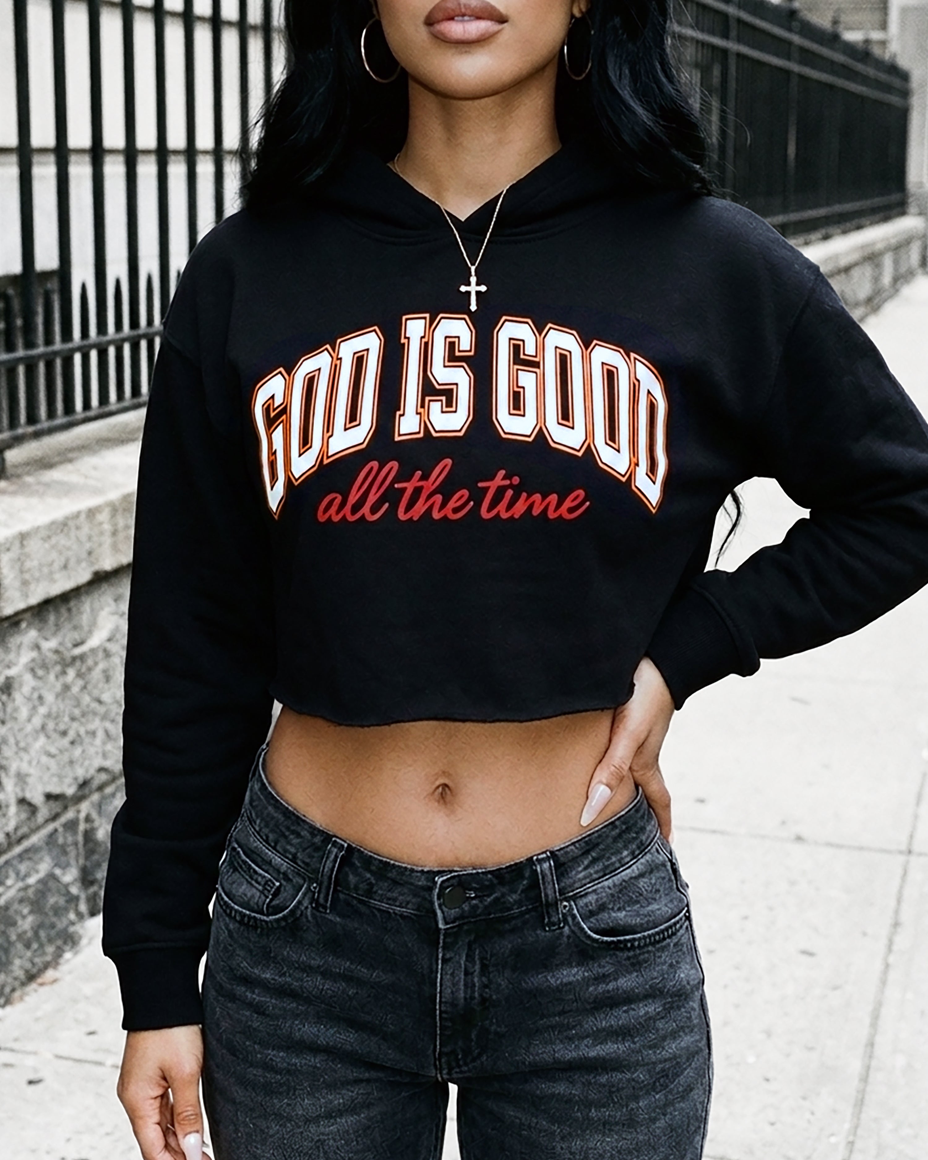 A woman wearing a black cropped hoodie with the text 'GOD IS GOOD all the time' in white and red, paired with dark gray jeans, standing outdoors on a sidewalk.
