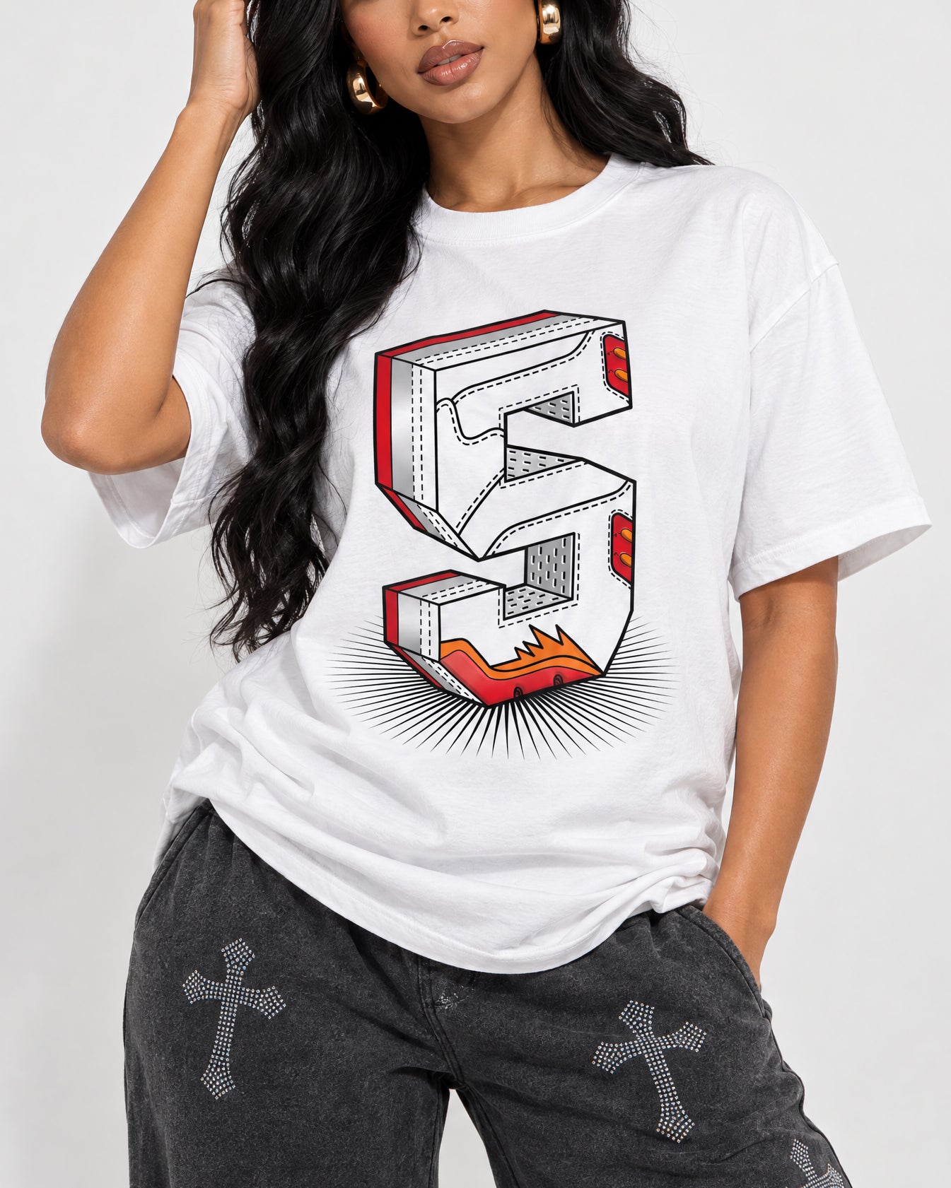 A woman wearing a white graphic t-shirt with a large 3D-style number 5 print in red, orange, and black, paired with dark gray jeans featuring cross details.
