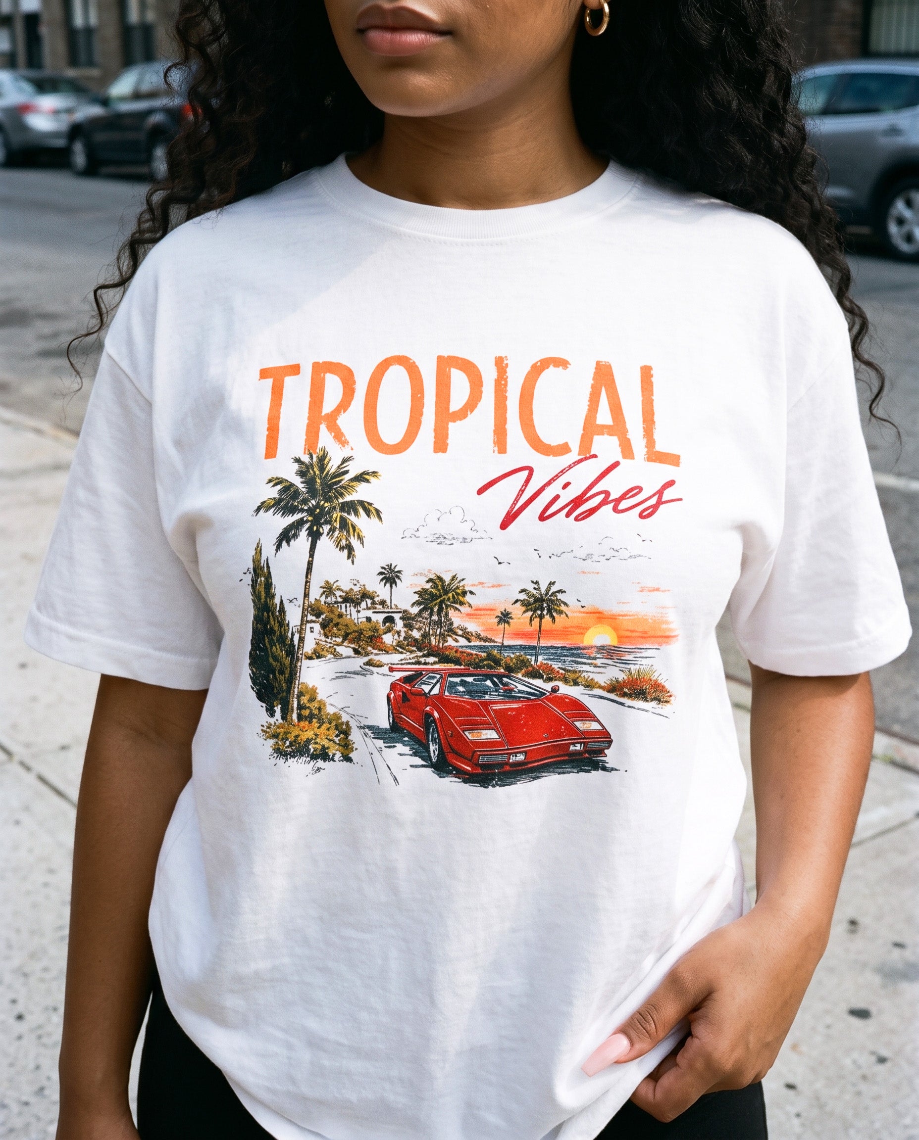 A woman wearing a white graphic t-shirt with 'TROPICAL Vibes' printed in orange and red, featuring a red sports car and palm trees on a beach sunset scene.