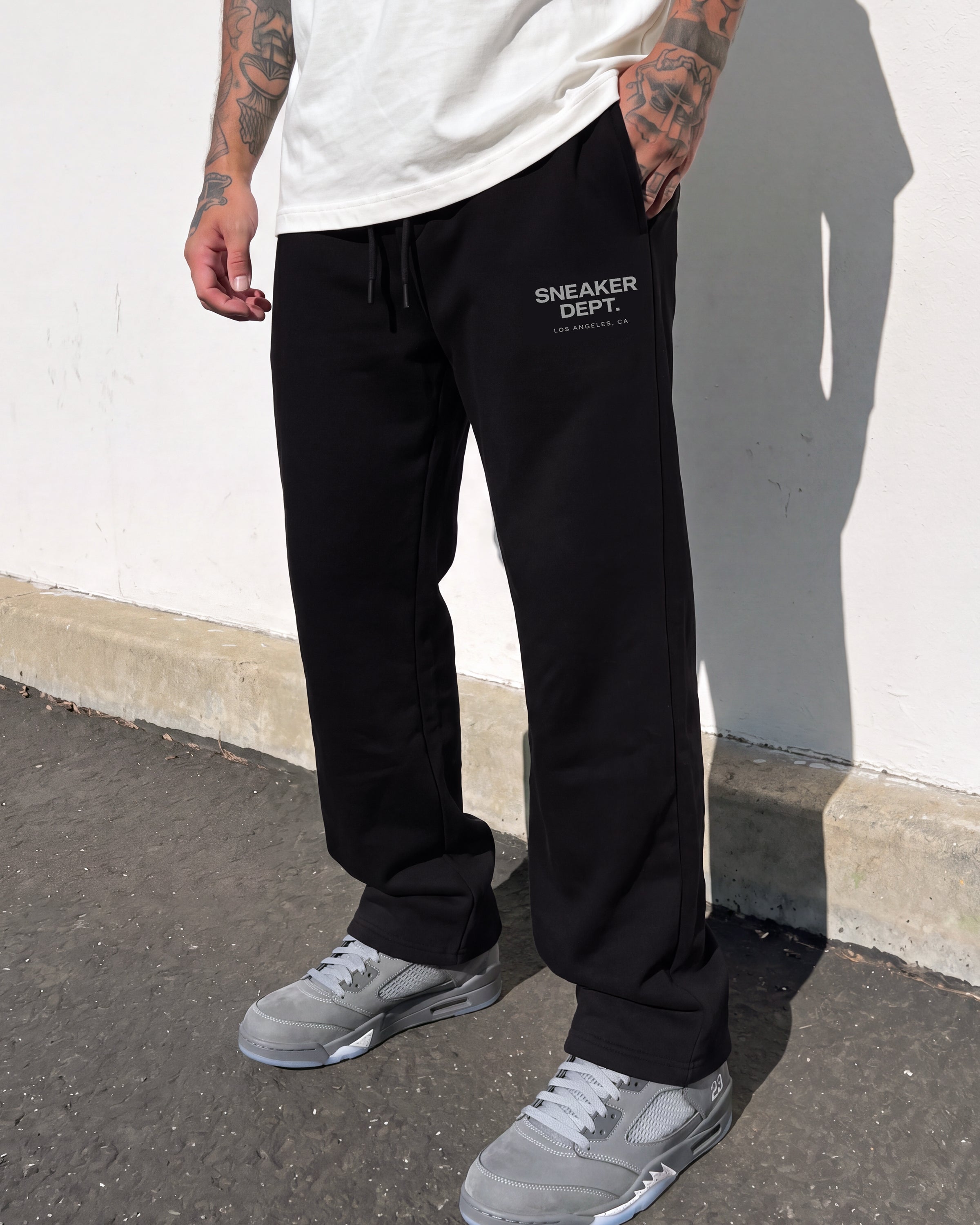 A person wearing black sweatpants with white 'SNEAKER DEPT.' text on the left thigh, paired with a white t-shirt and gray sneakers, standing outdoors against a white wall.