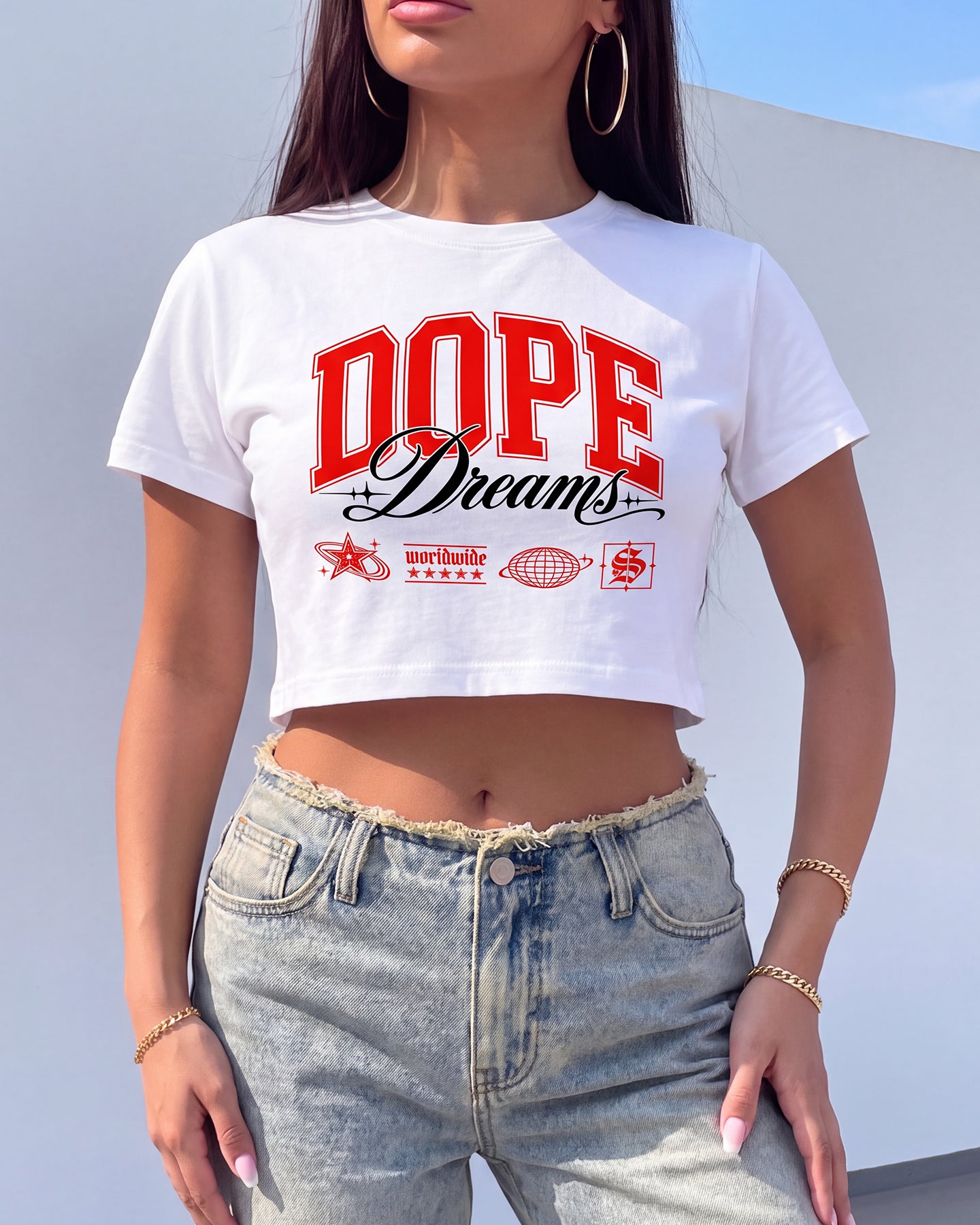 A woman wearing a white cropped t-shirt with a red and black 'DOPE Dreams' graphic, paired with light-wash frayed-hem jeans, set against a minimalist light gray wall background.