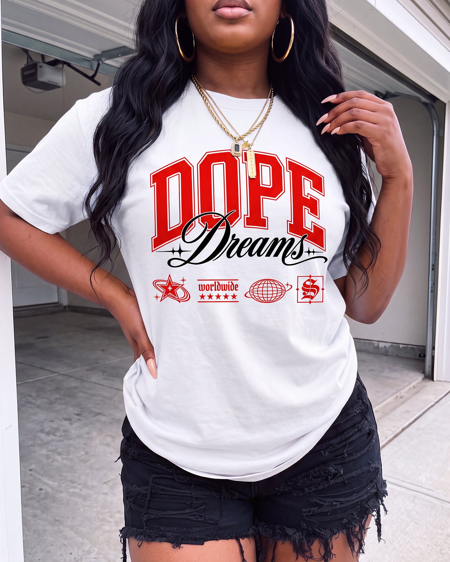 A woman wearing a white graphic t-shirt with 'DOPE Dreams' printed in red and black, paired with black distressed denim shorts, standing in front of a garage door.