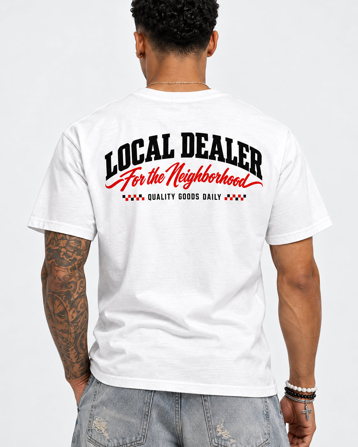 A man viewed from behind wearing a white t-shirt with a graphic on the back that reads 'LOCAL DEALER For the Neighborhood' in black and red, paired with light-wash distressed jeans against a light gray background.
