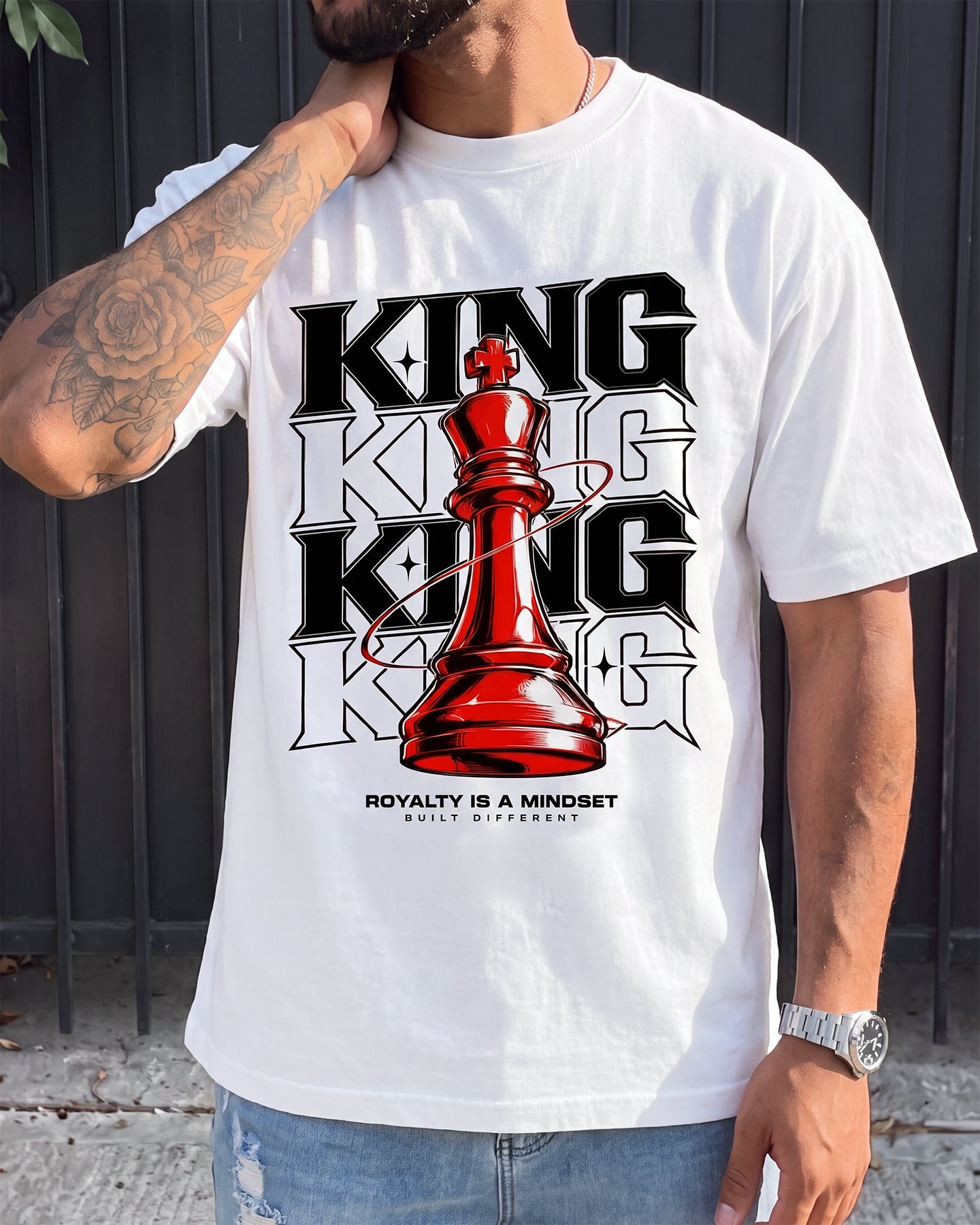 A man wearing a white graphic t-shirt with a bold black and red design featuring the word 'KING' and a red chess piece, paired with blue jeans, set against a dark metal fence background.
