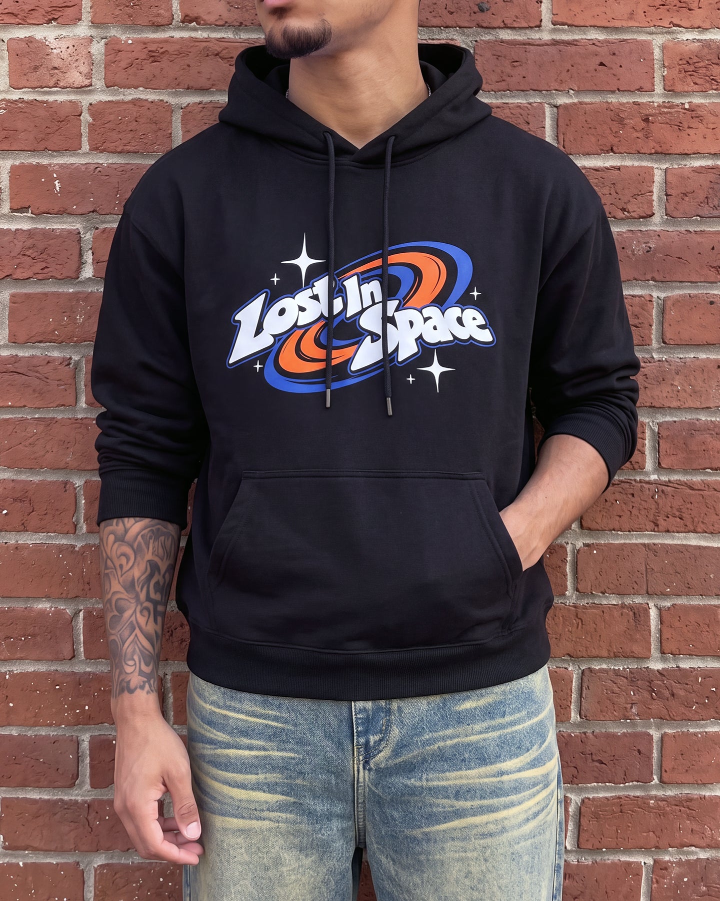 A man wearing a black pullover hoodie with a colorful 'Lost in Space' graphic on the chest, paired with light-wash distressed jeans, standing against a red brick wall.