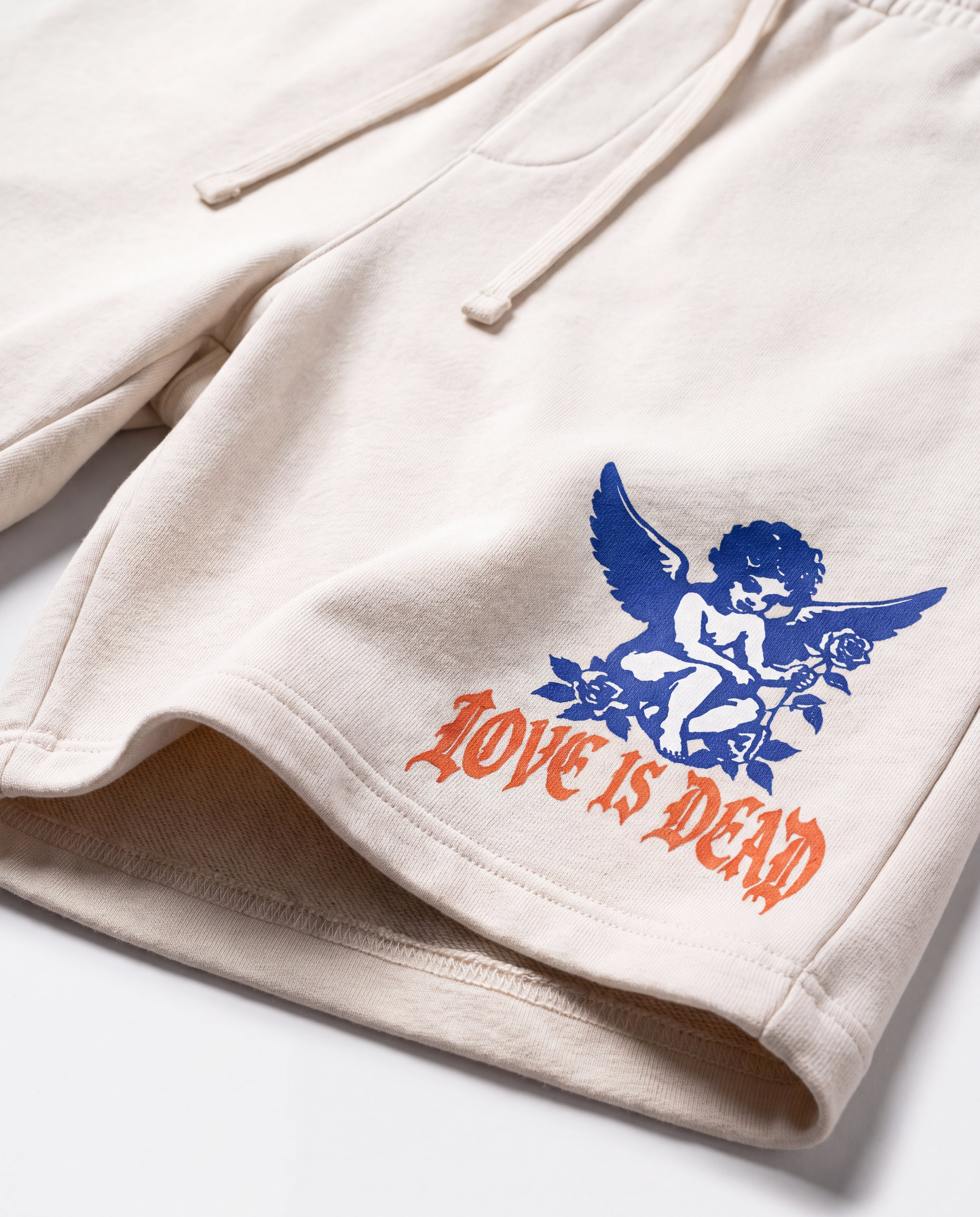 Close-up of cream-colored drawstring shorts with a blue cherub graphic and orange gothic text reading 'LOVE IS DEAD' on the lower left leg.