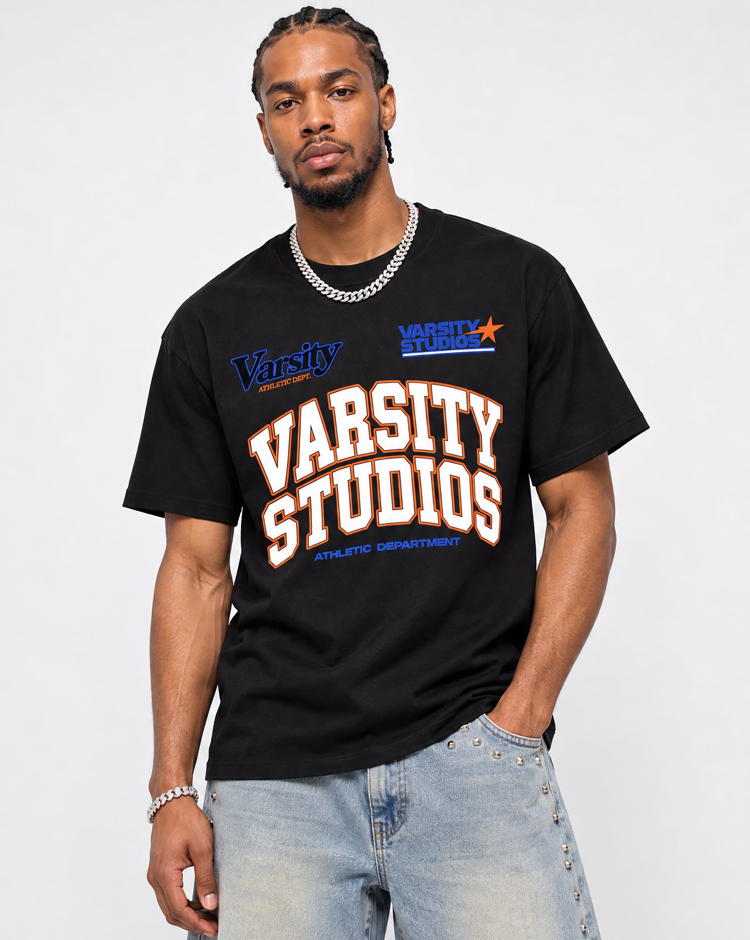 A man wearing a black graphic t-shirt with 'Varsity Studios' printed in white and orange, paired with light blue jeans and a silver chain necklace, against a plain light gray background.