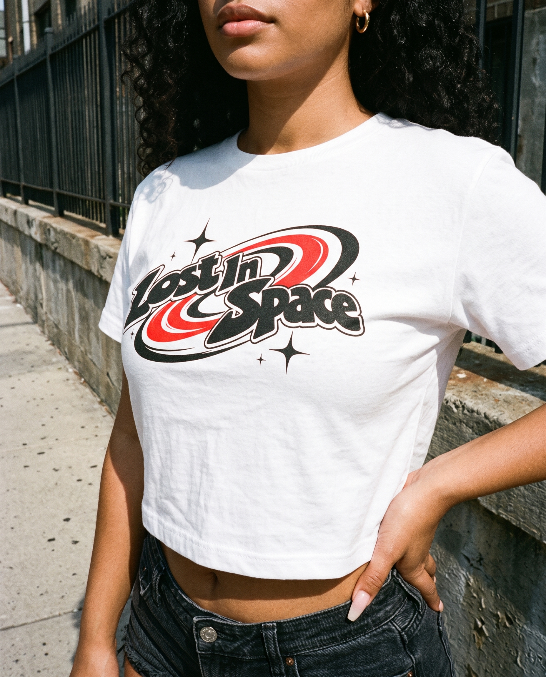 A woman wearing a white cropped t-shirt with a red, black, and white 'Lost in Space' graphic on the front, paired with dark denim shorts, standing outdoors next to a stone wall.