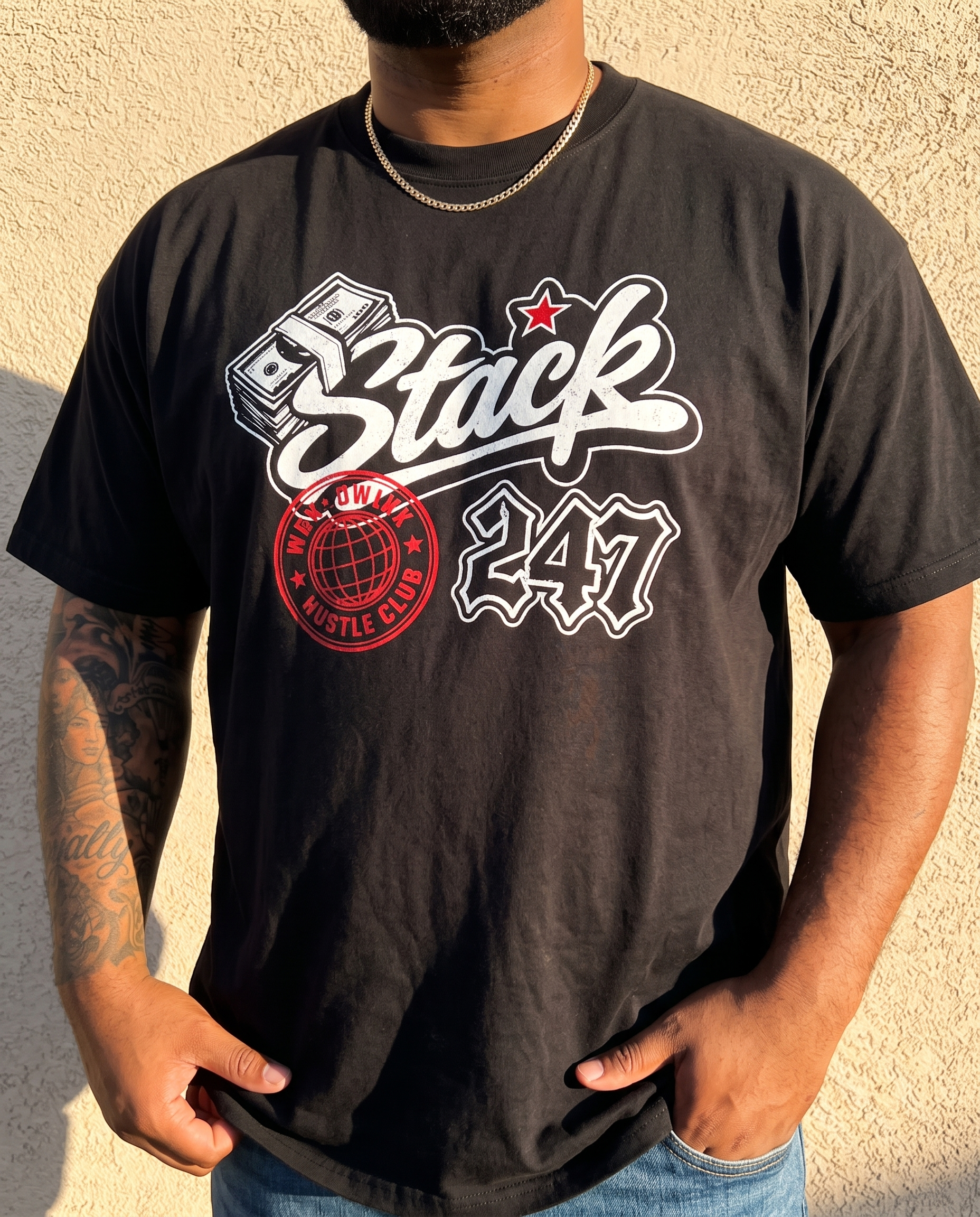 A man wearing a black graphic t-shirt with a white and red 'Stack 247' design, standing against a textured beige wall.